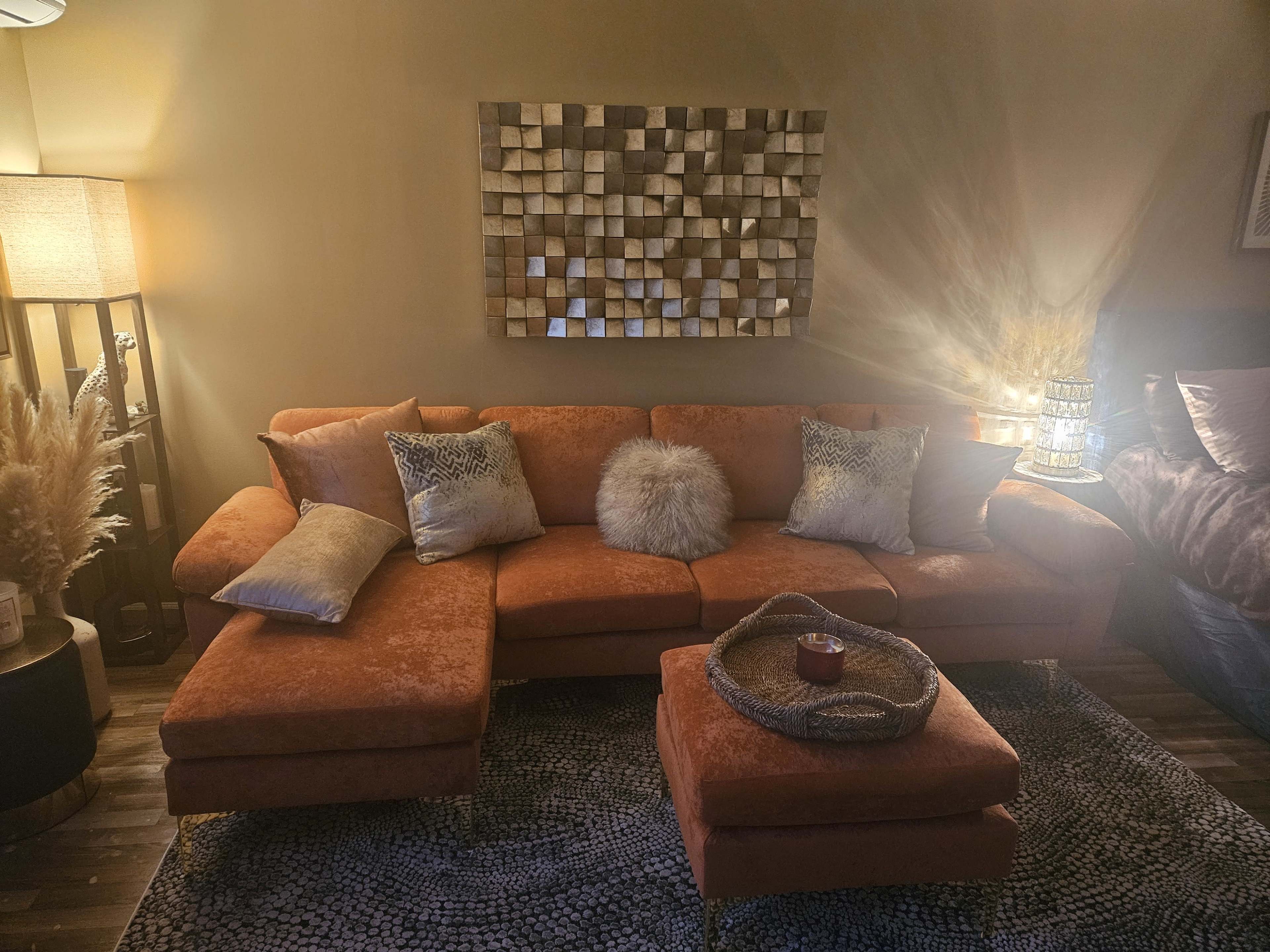 A cozy living room features an orange sectional sofa with decorative pillows, a lighted lamp, and a wall adorned with a geometric mirror above a textured rug.