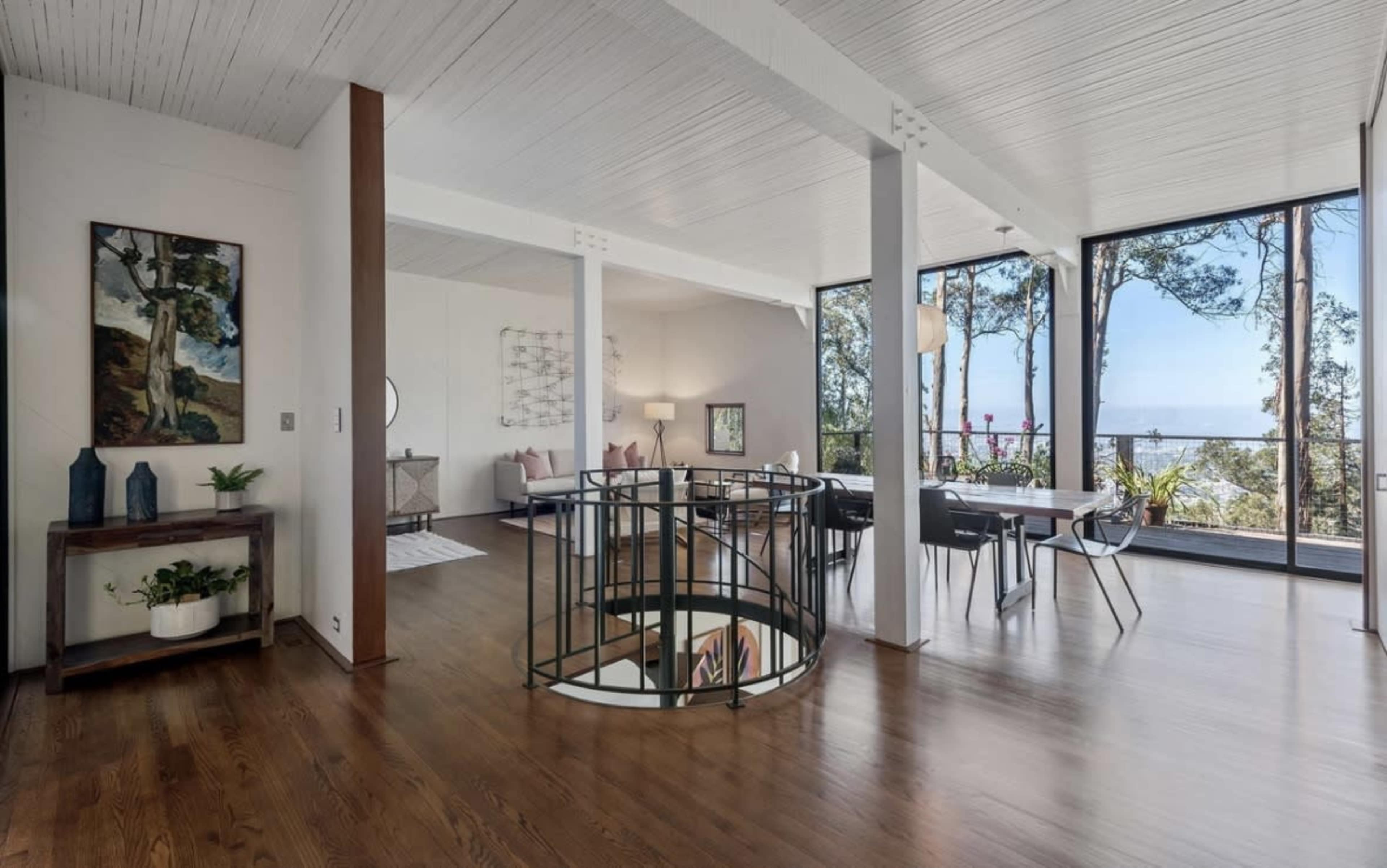A modern, open-concept living area with large windows providing views of trees and a body of water, featuring wooden flooring and a curved staircase.
