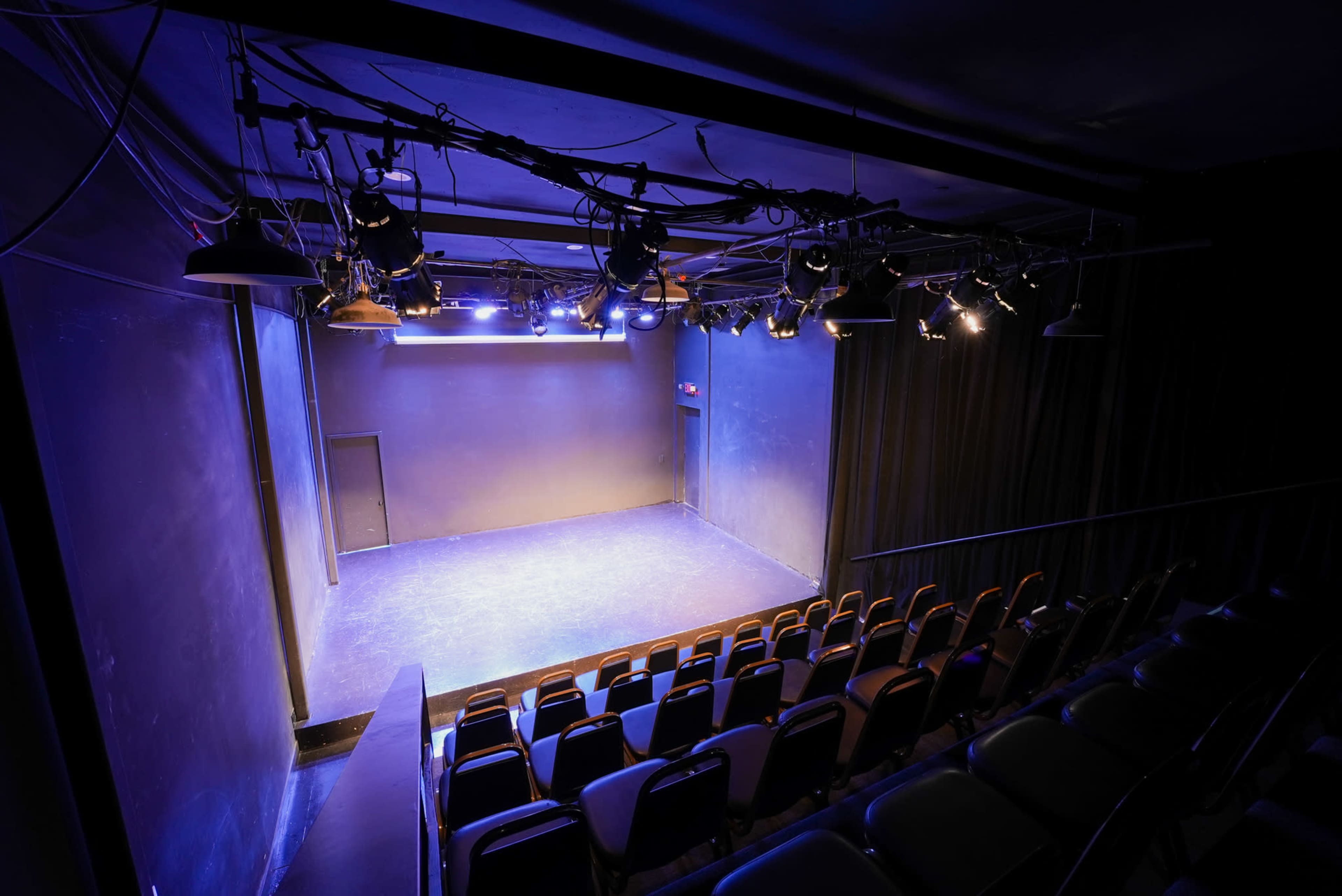 The image shows an empty theater stage with blue lighting and rows of seats visible in the background.