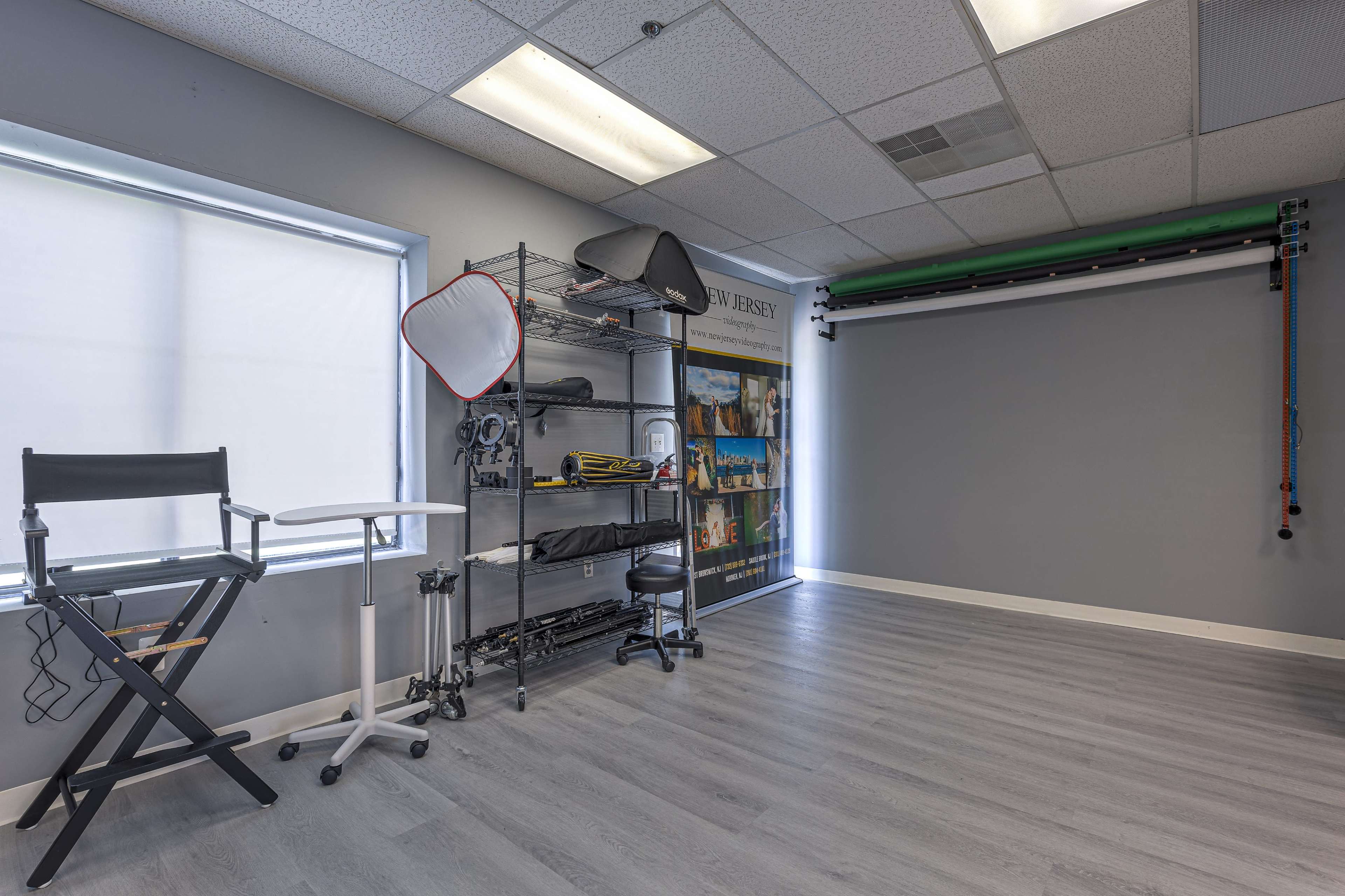 A spacious photography studio with a gray wall, a director's chair, a mobile cart, and various photography equipment neatly arranged on a shelving unit.