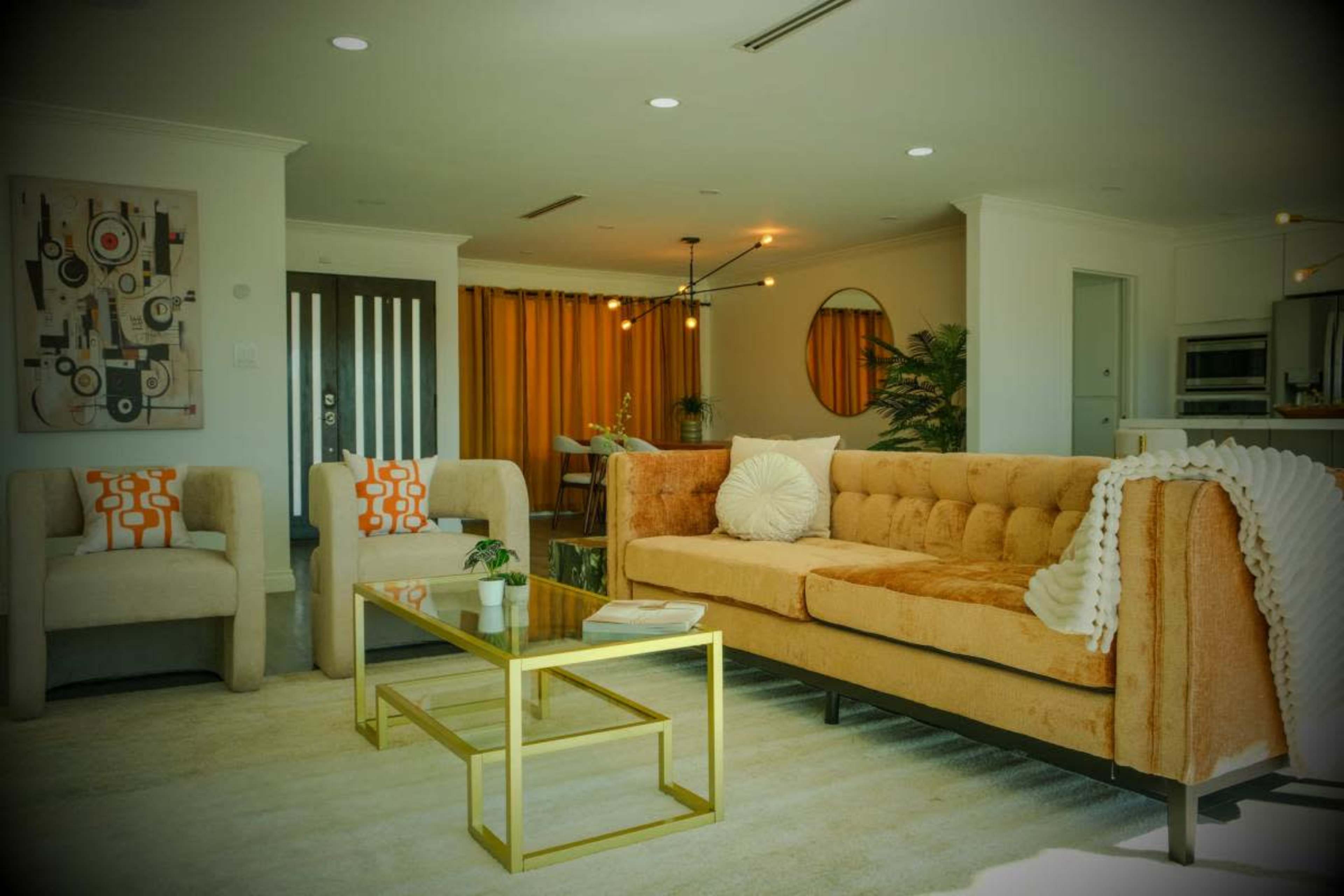 The image shows a modern living room with an orange couch, two light-colored chairs, a glass coffee table, and large windows covered by yellow curtains.