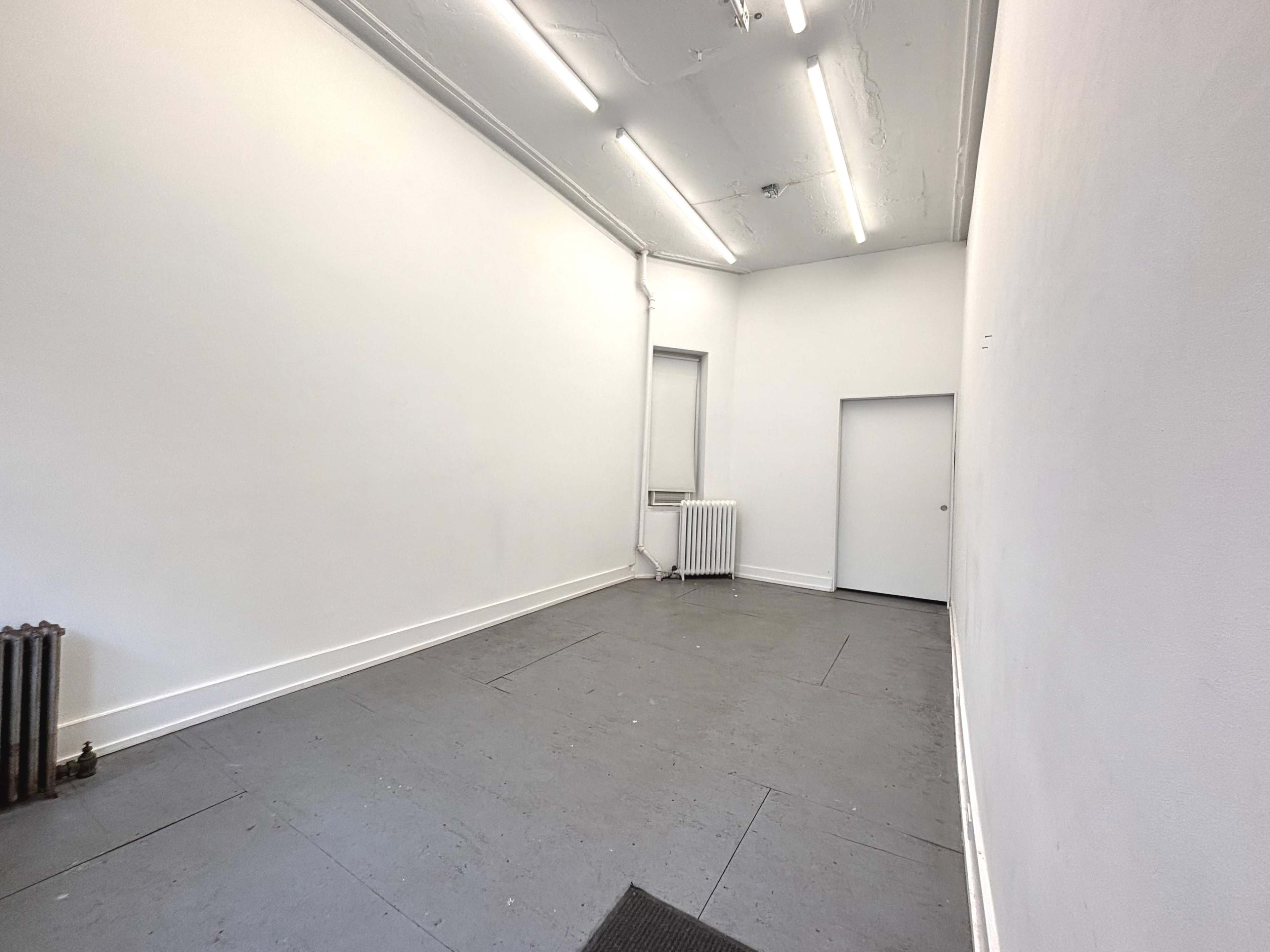 The image shows a vacant, white-walled room with a gray floor, a radiator in the corner, and a door at one end.