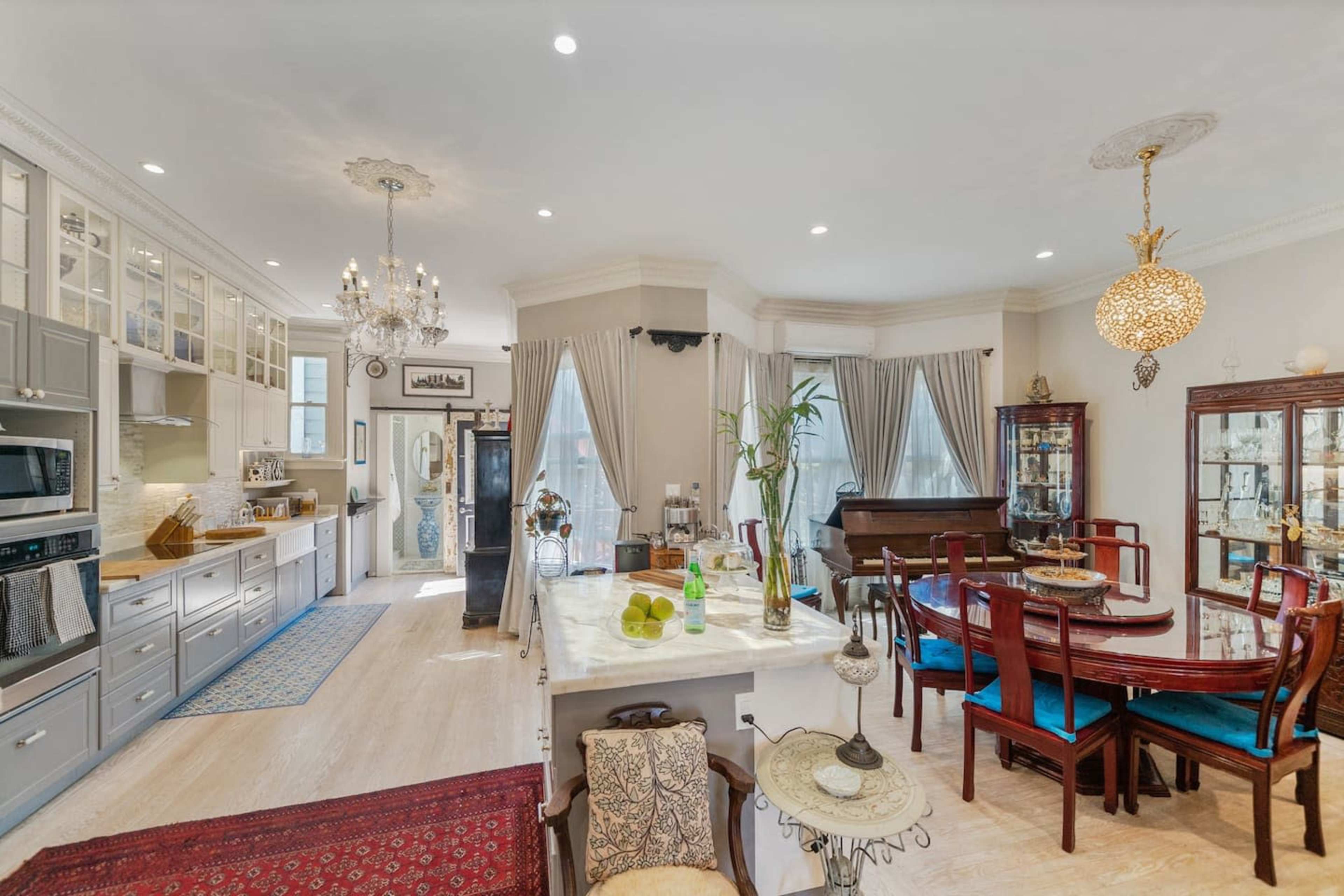 A bright kitchen and dining area features a central table surrounded by chairs, cabinetry, and a piano in a spacious layout.