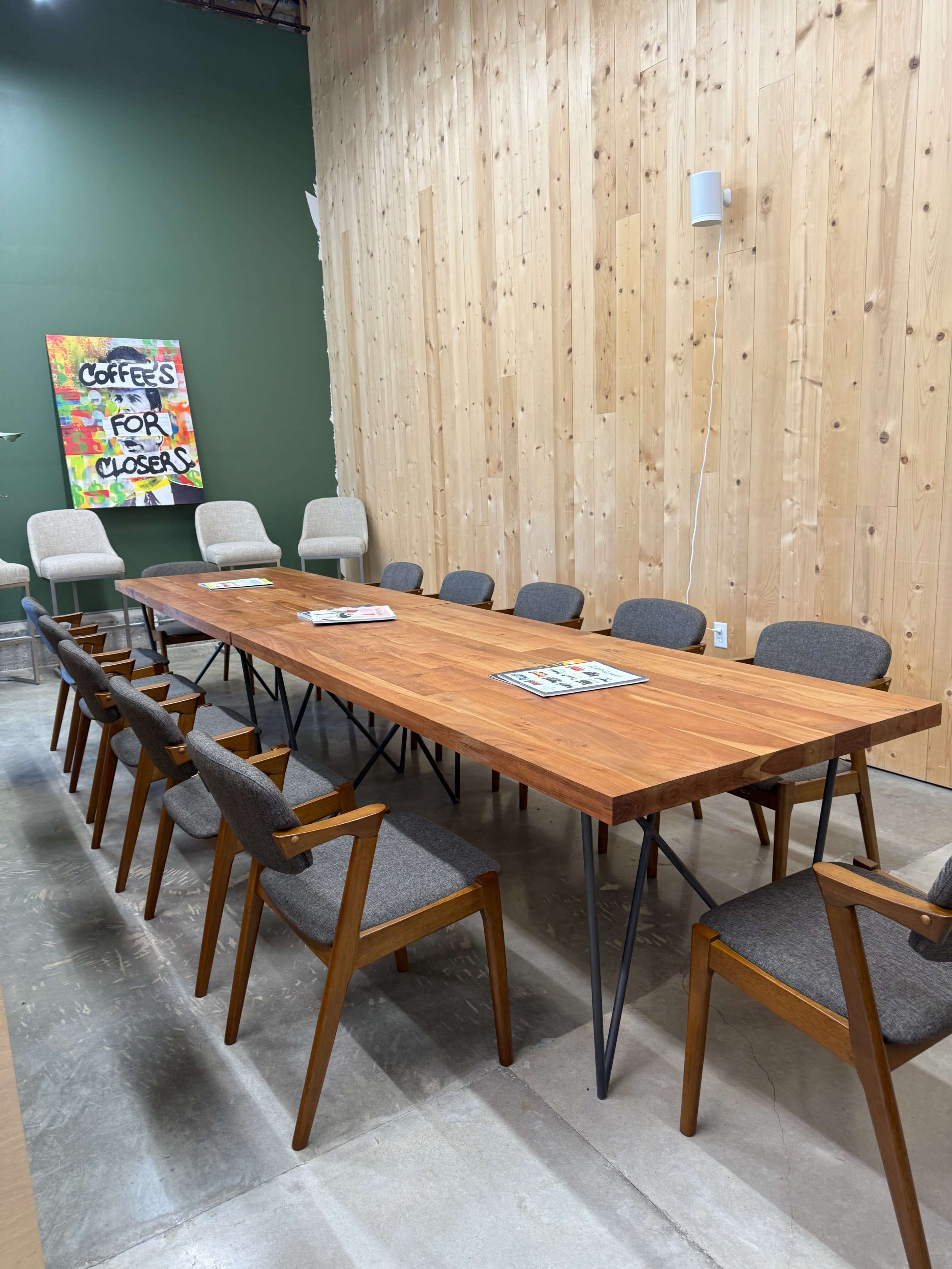 A large wooden conference table surrounded by several chairs is set against a wooden wall, with a colorful artwork saying "Coffee’s for Closers" in the background.