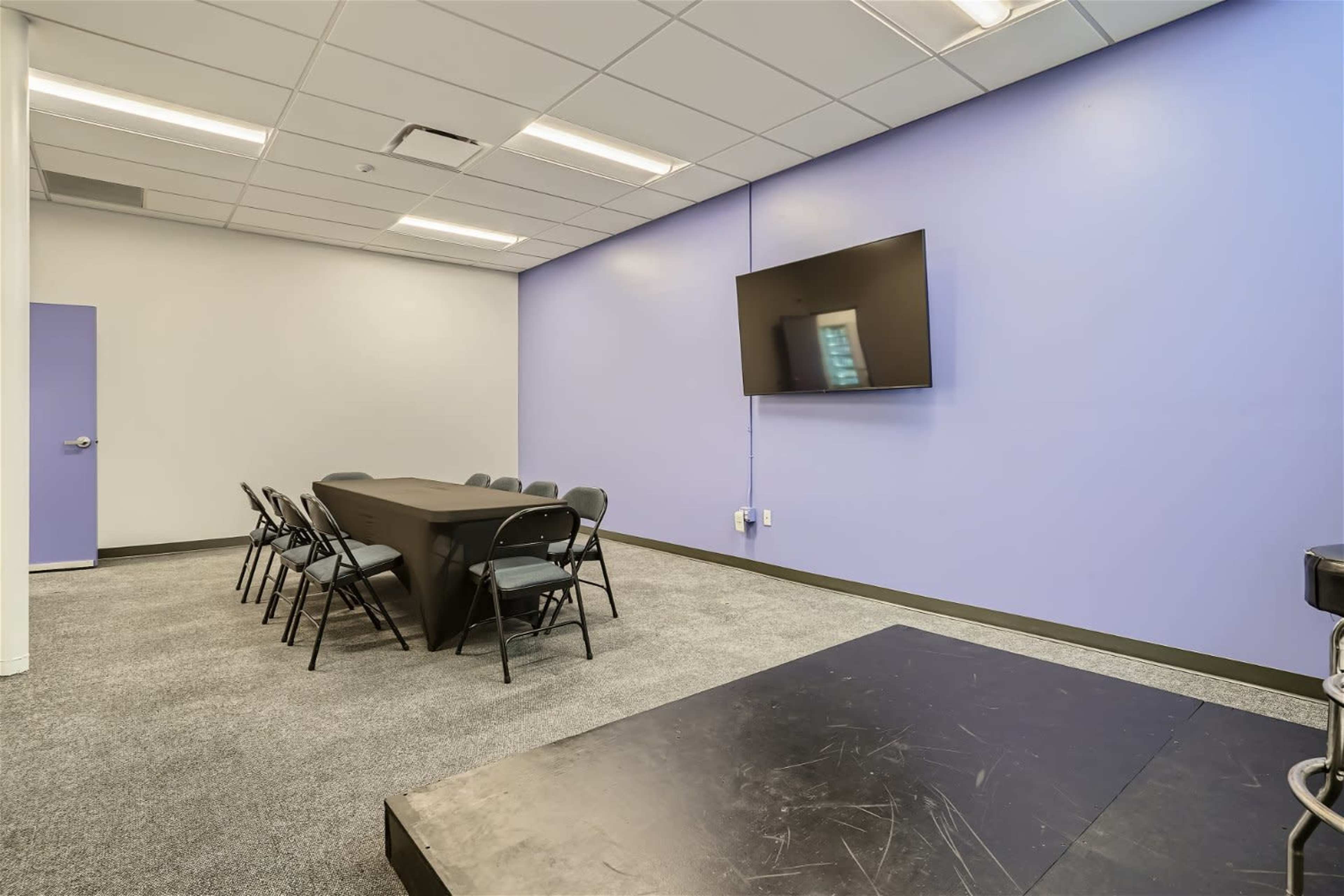 A meeting room features a black table surrounded by seven folding chairs, a large flat-screen television mounted on a purple wall, and gray carpeting.