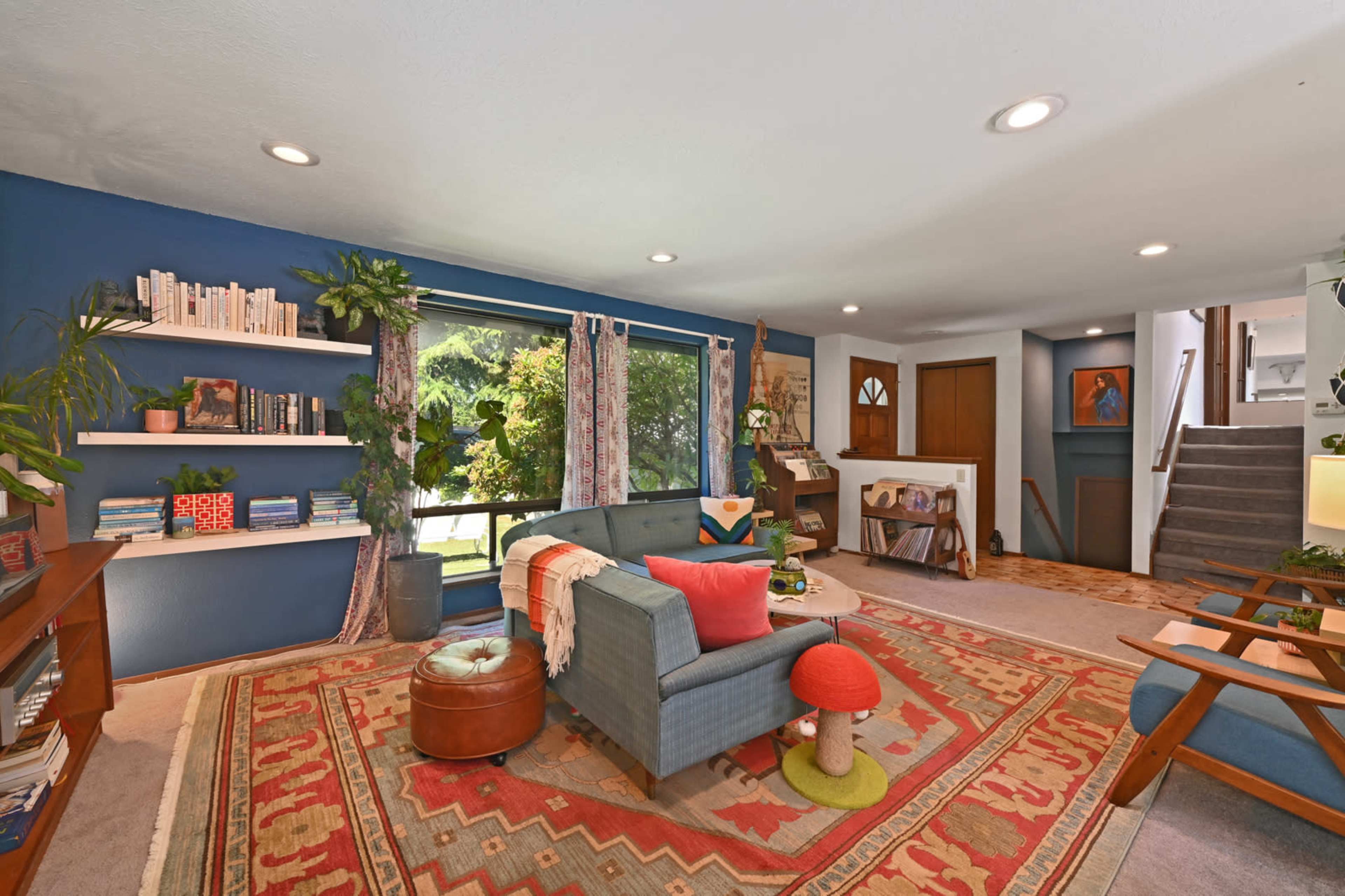 The image shows a living room with a blue accent wall, a large window displaying greenery outside, a sectional sofa, a patterned area rug, and various bookshelves.