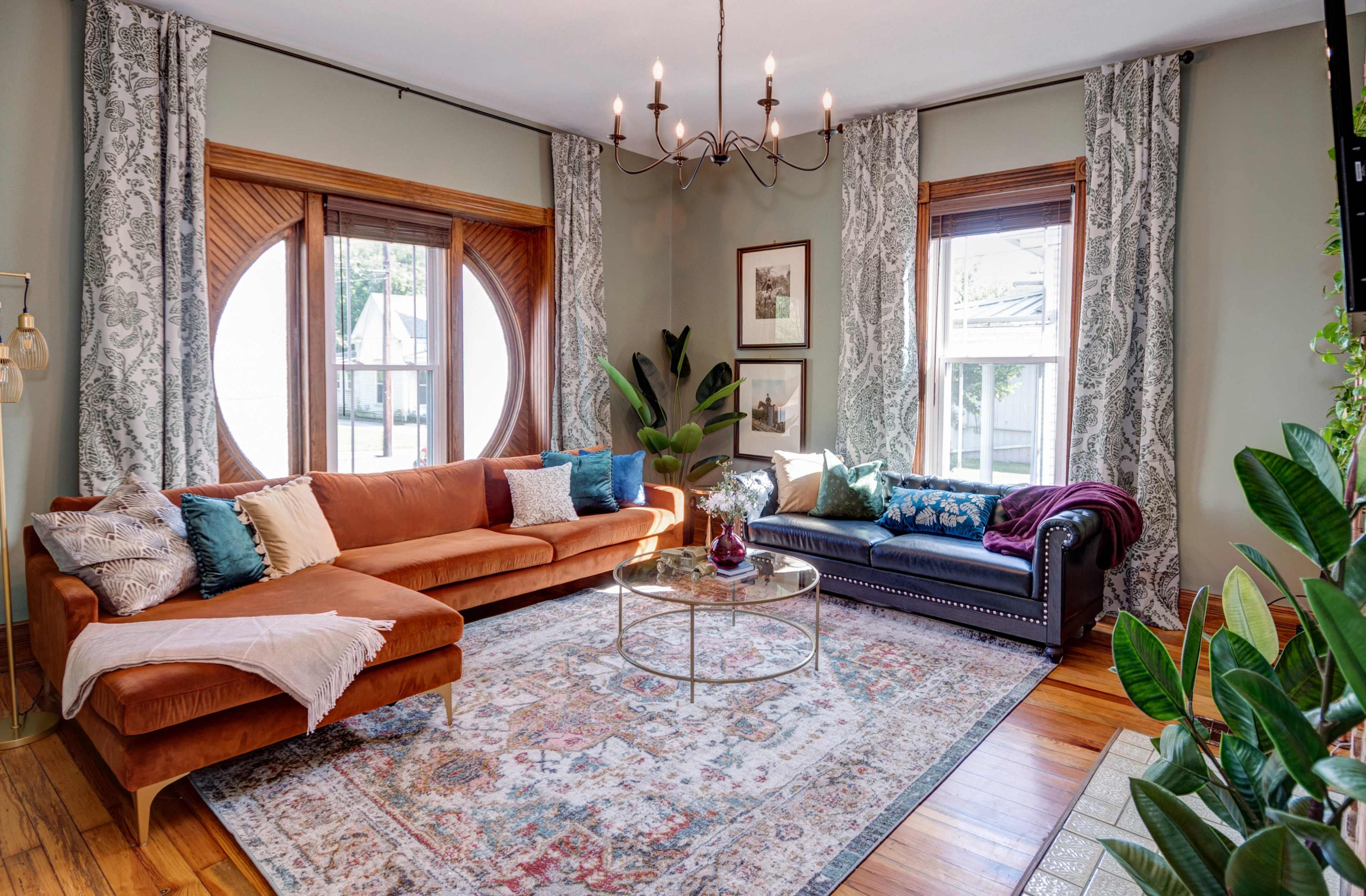A living room with a brown sectional sofa, a black sofa, a round coffee table on a patterned rug, and large windows with decorative curtains.