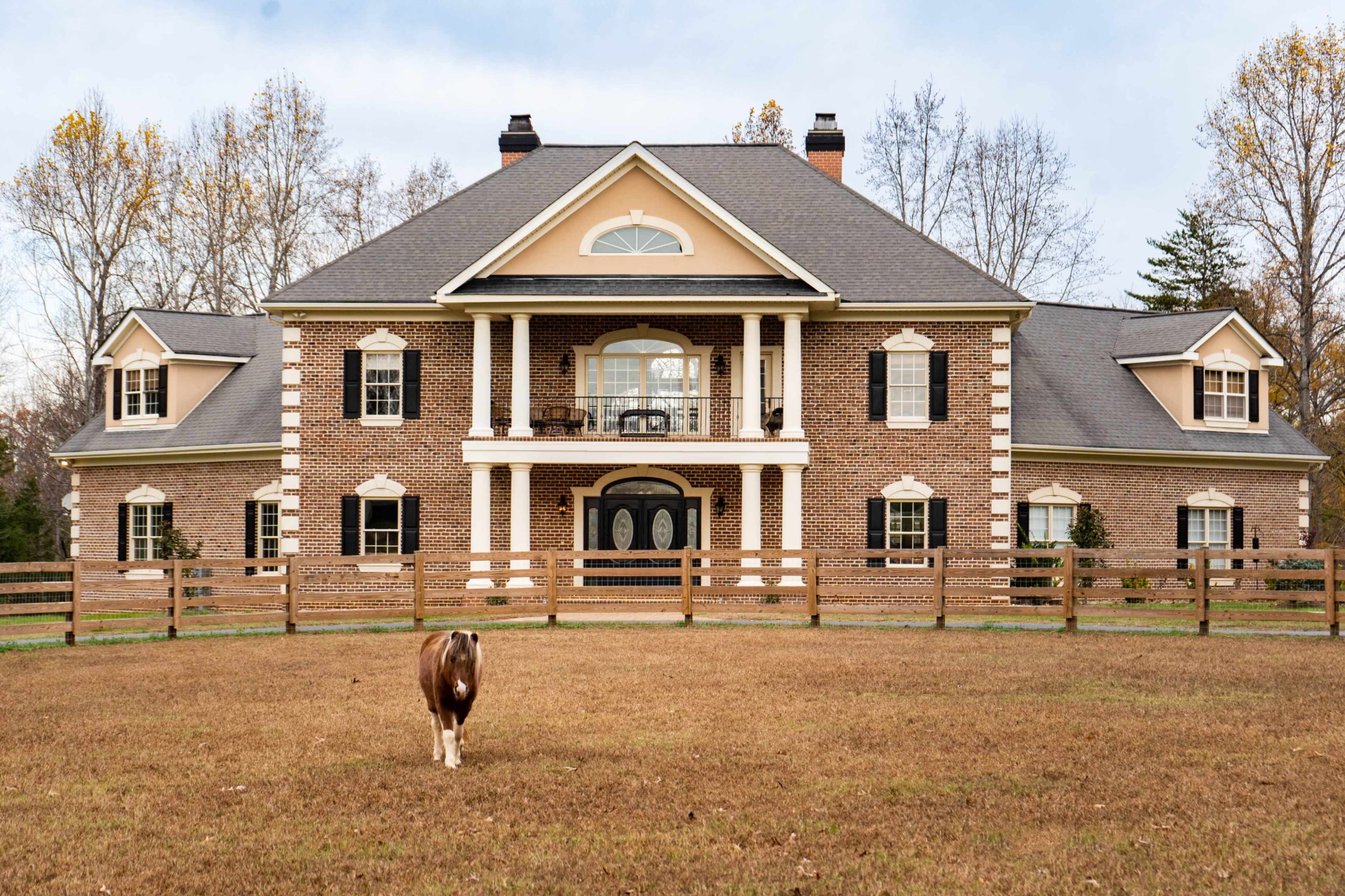 A large brick house with multiple gables and a front porch stands in a grassy field, while a brown and white cow grazes nearby.
