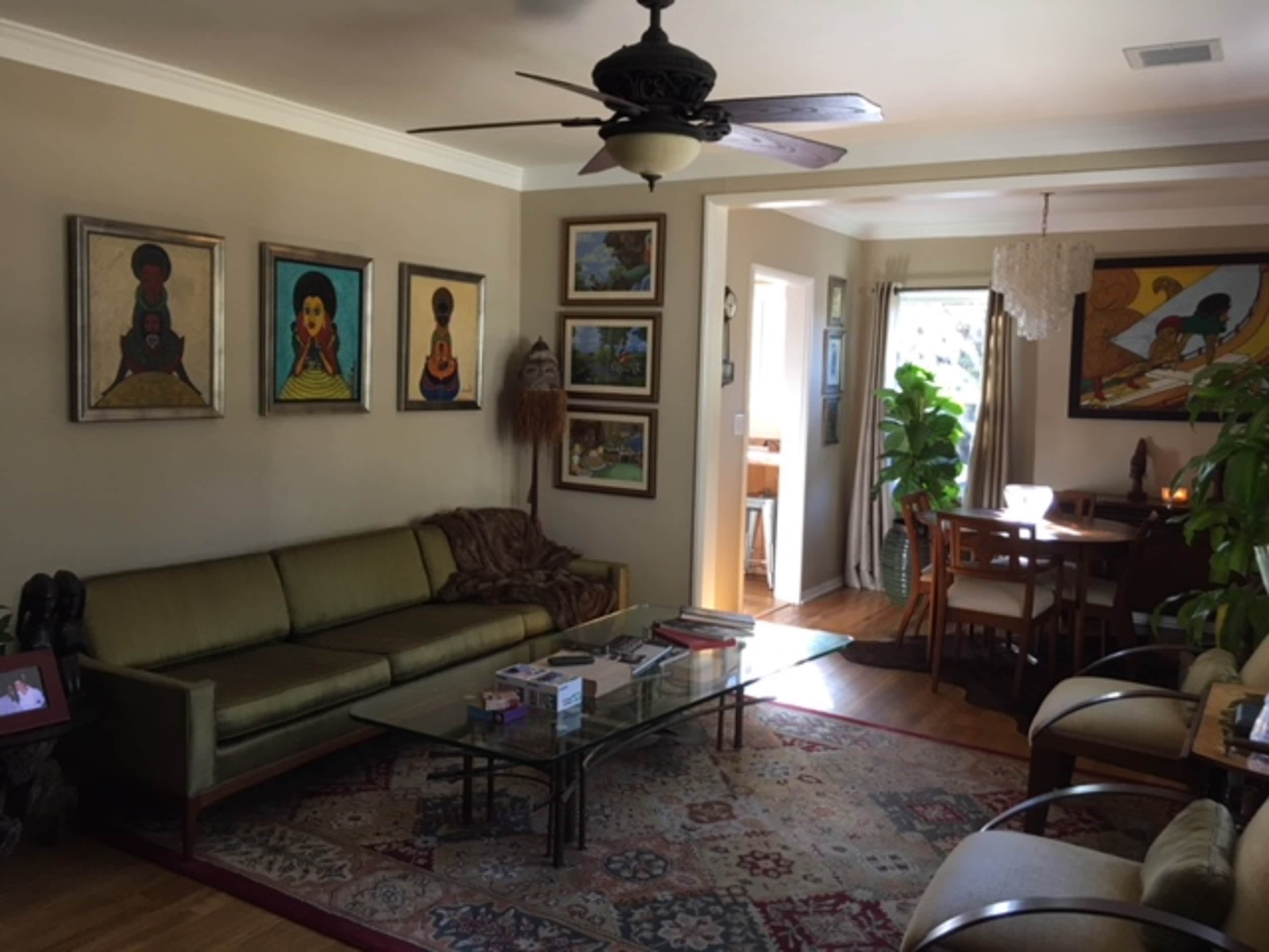 The living room features a green sofa, a glass coffee table, and several framed artworks on the walls, with a dining area visible in the background.
