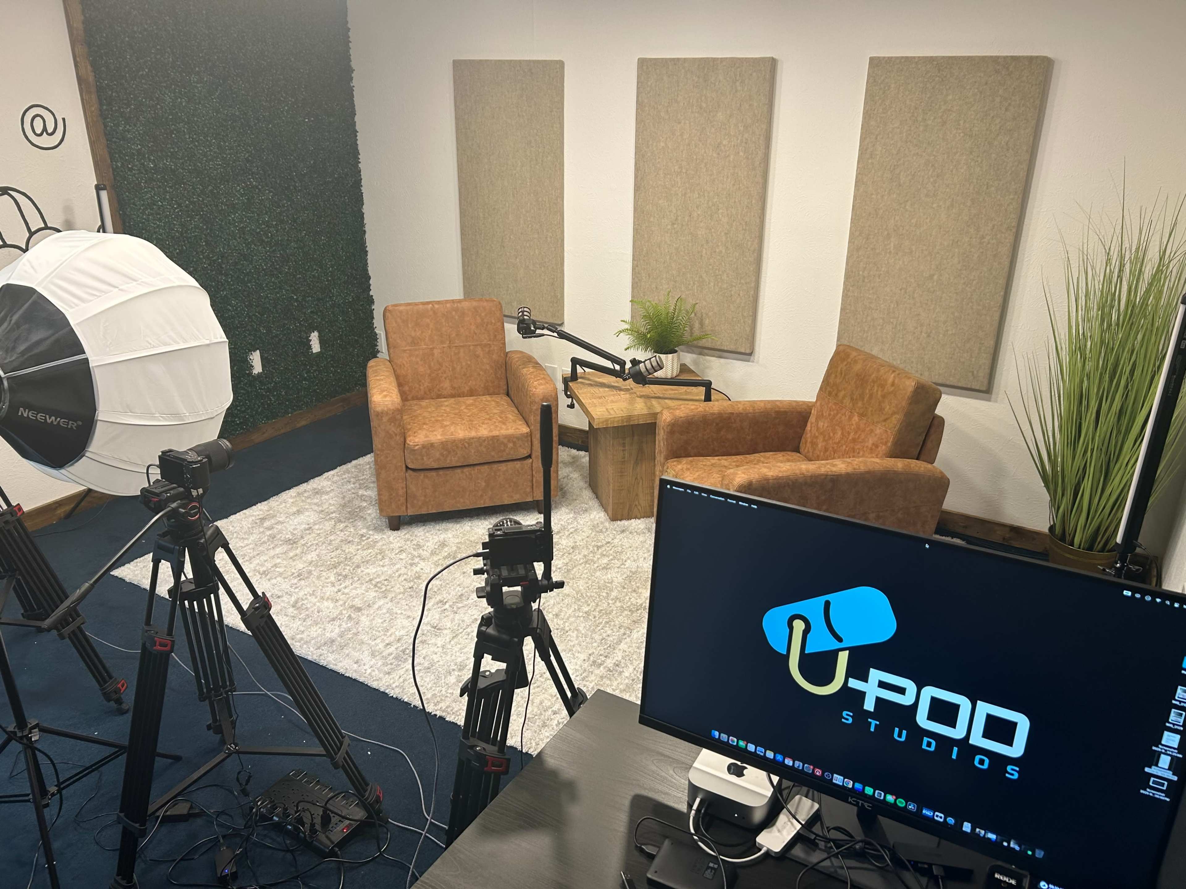 The image shows a recording studio setup with two brown chairs, a wooden table, a large softbox light, several cameras on tripods, and a computer displaying the logo of "U-Pod Studios."