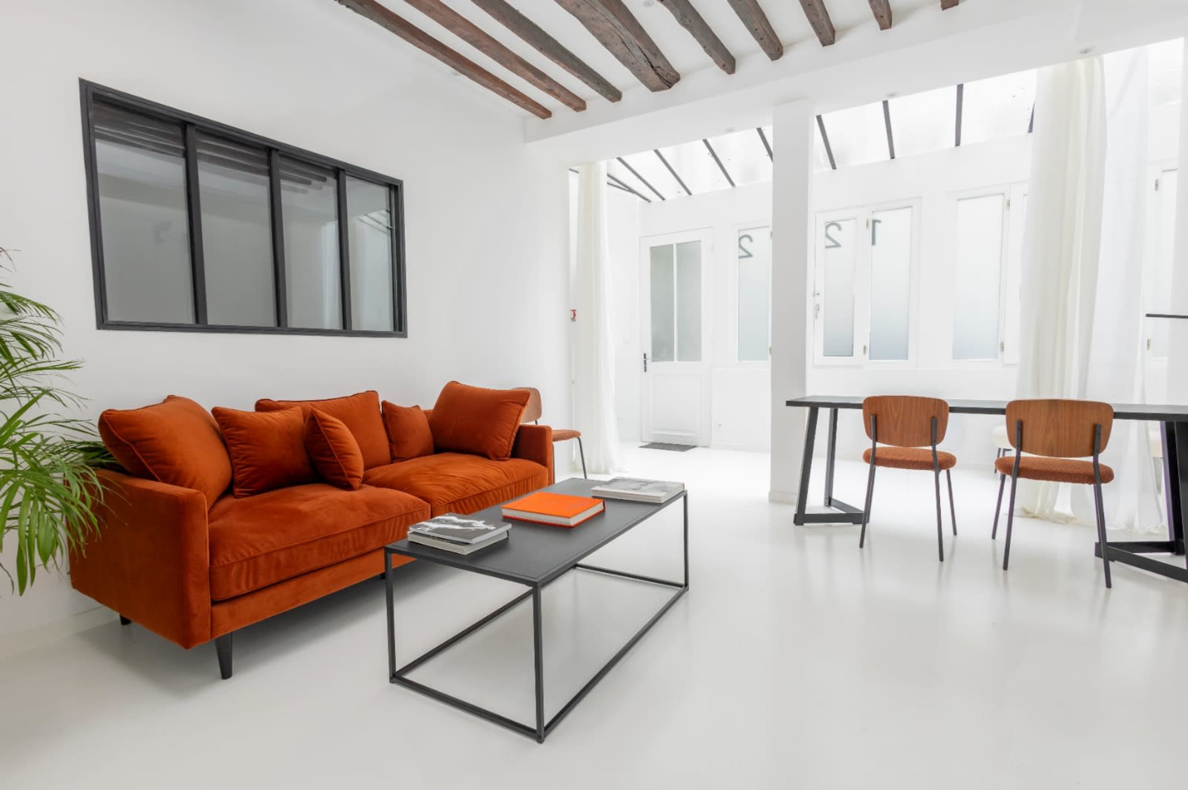 The image shows a bright, modern living space featuring an orange sofa, a glass coffee table, and a dining area with wooden chairs.