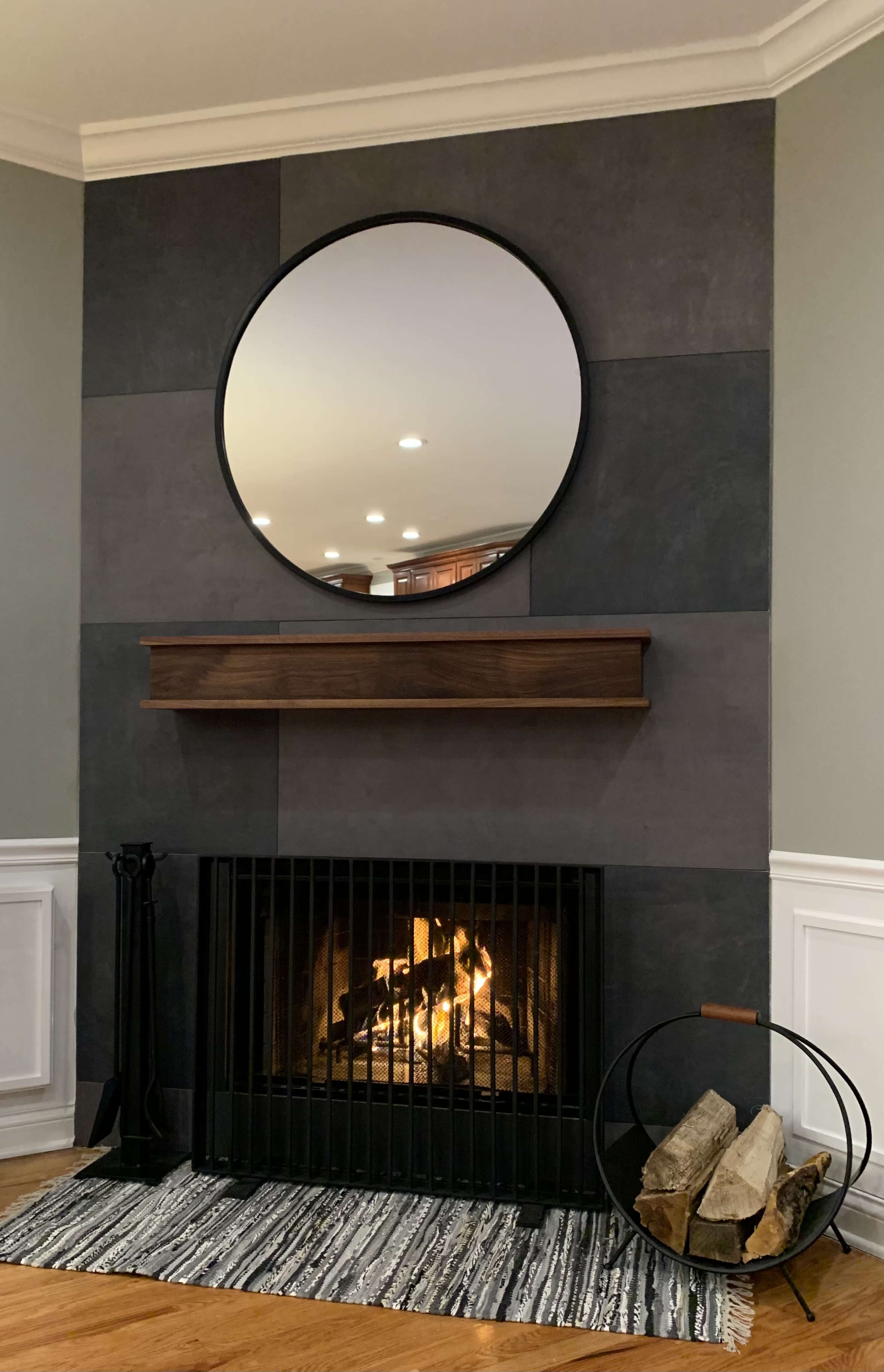 A round mirror hangs above a wooden mantel positioned over a fireplace, which has a fire burning, and logs are neatly stacked beside it on a patterned rug.