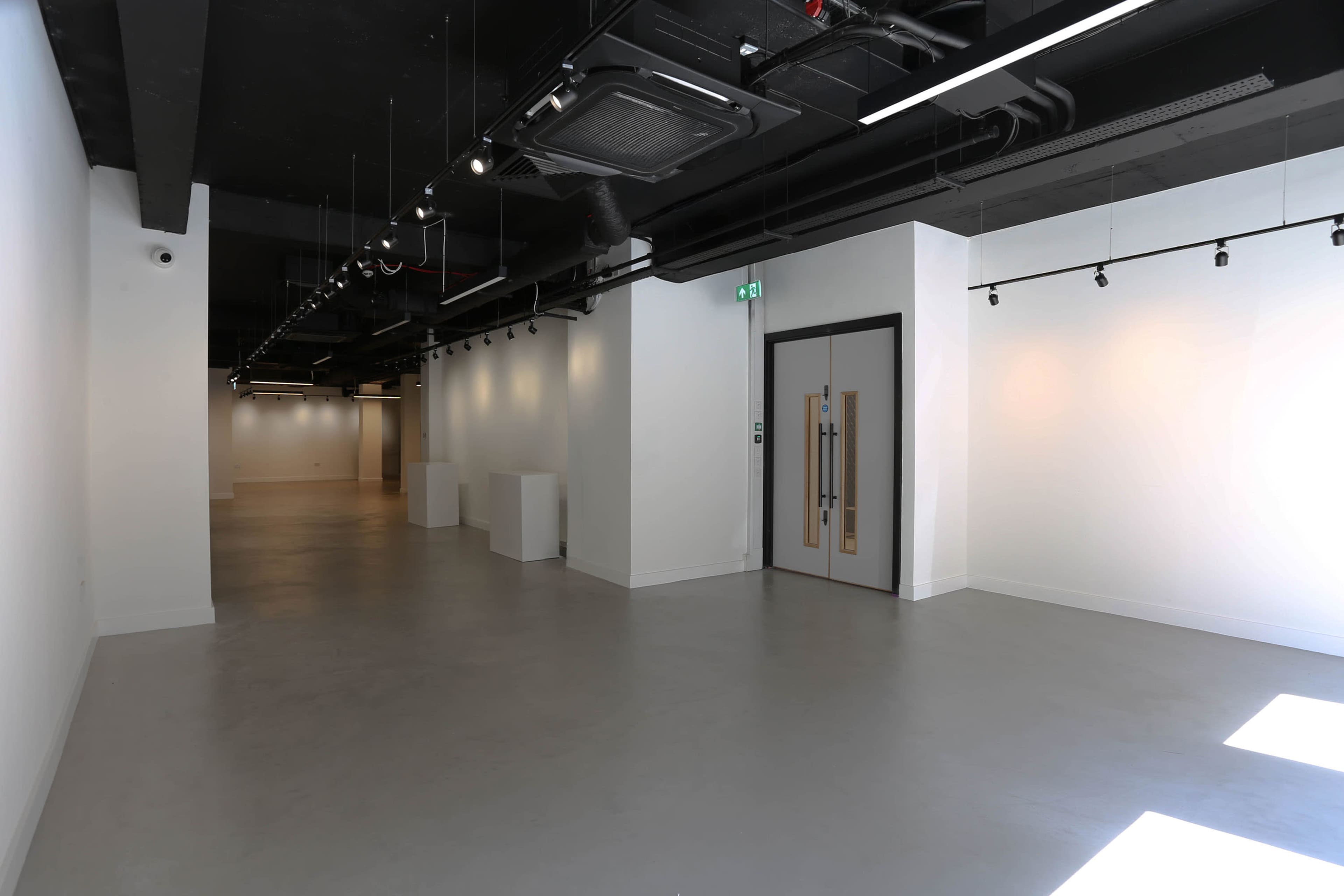 The image shows a spacious, empty interior with white walls and a polished concrete floor, featuring overhead lighting and a double-door entrance at one end.