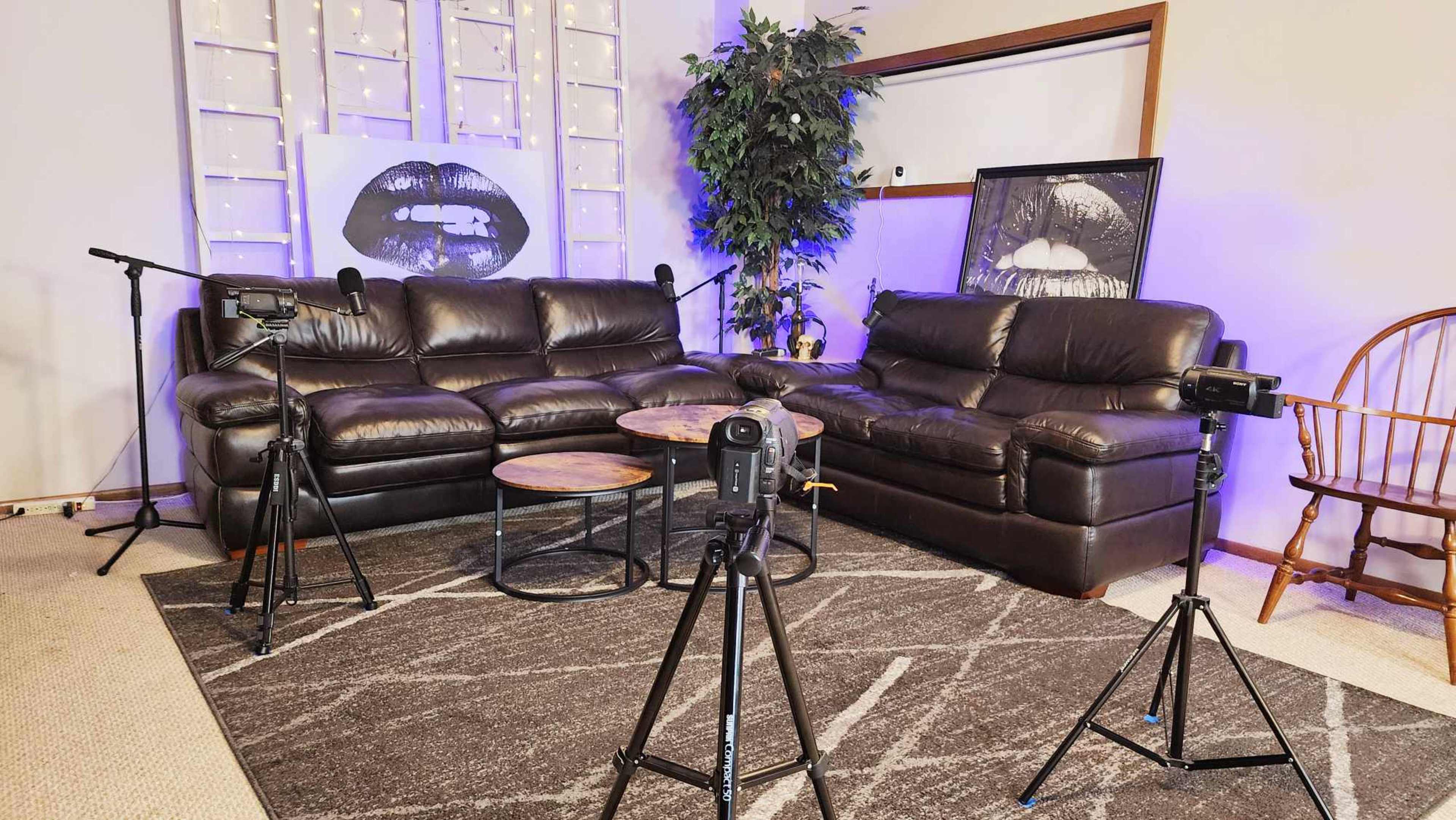 The scene features a comfortable living room setup with a dark leather sectional sofa, two cameras on tripods, a round wooden coffee table, and a decorative plant against a backdrop of illuminated wall art.