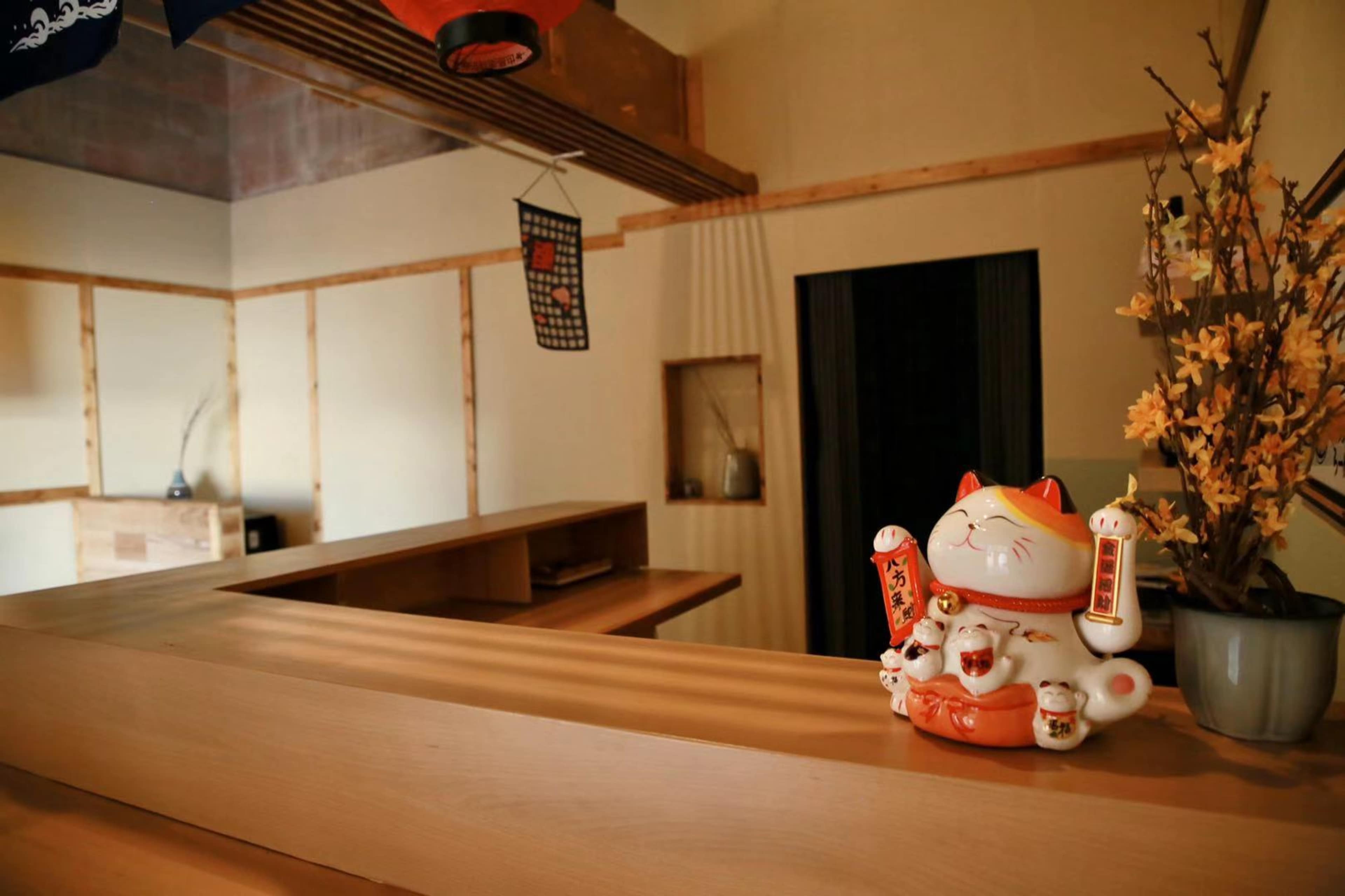 The image shows a traditional Japanese-style interior with a wooden counter, a fortune cat figurine, and decorative branches in a vase.