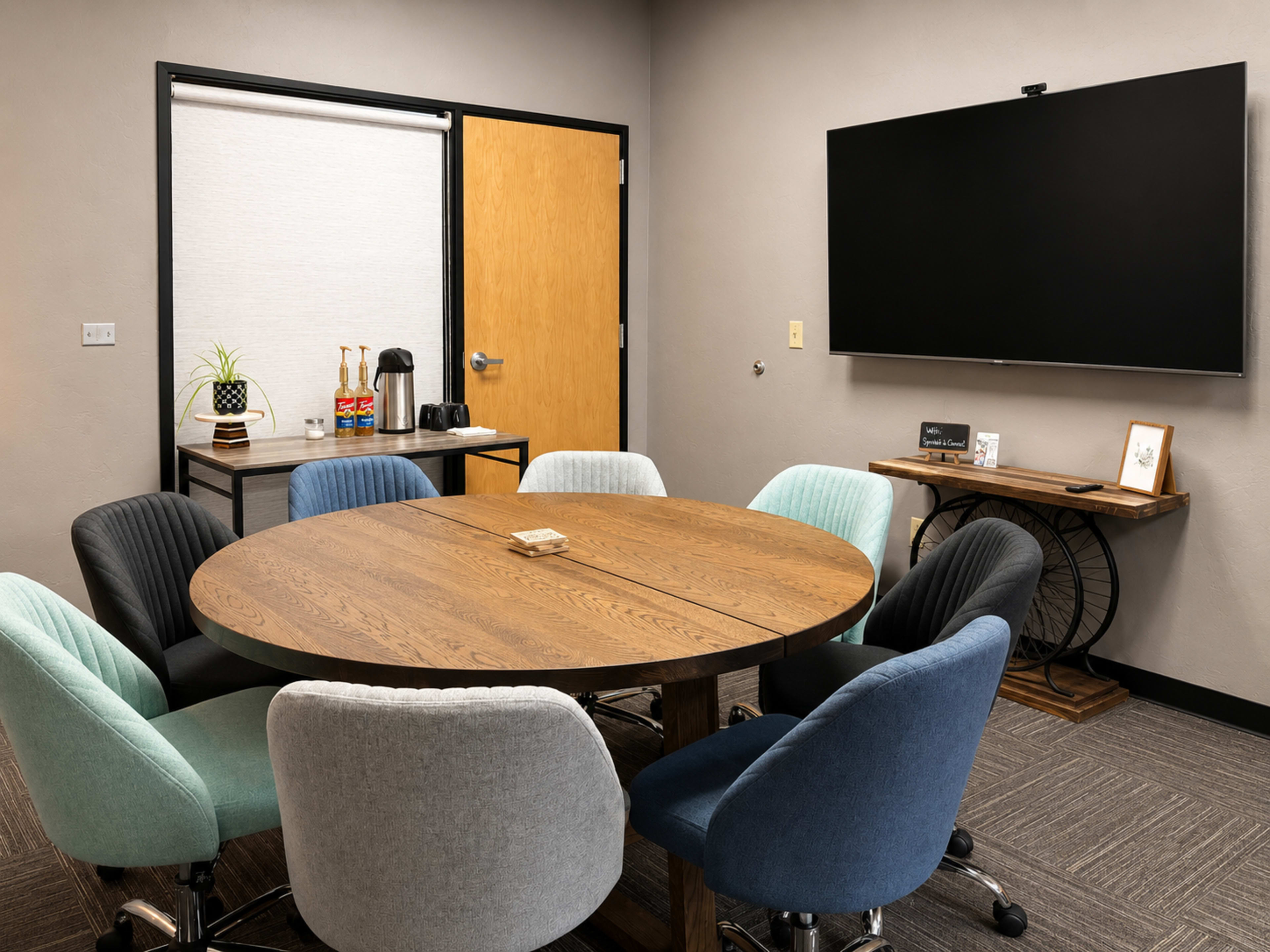 The image shows a meeting room featuring a large round wooden table surrounded by eight colorful upholstered chairs, with a television mounted on the wall and a coffee station in the corner.
