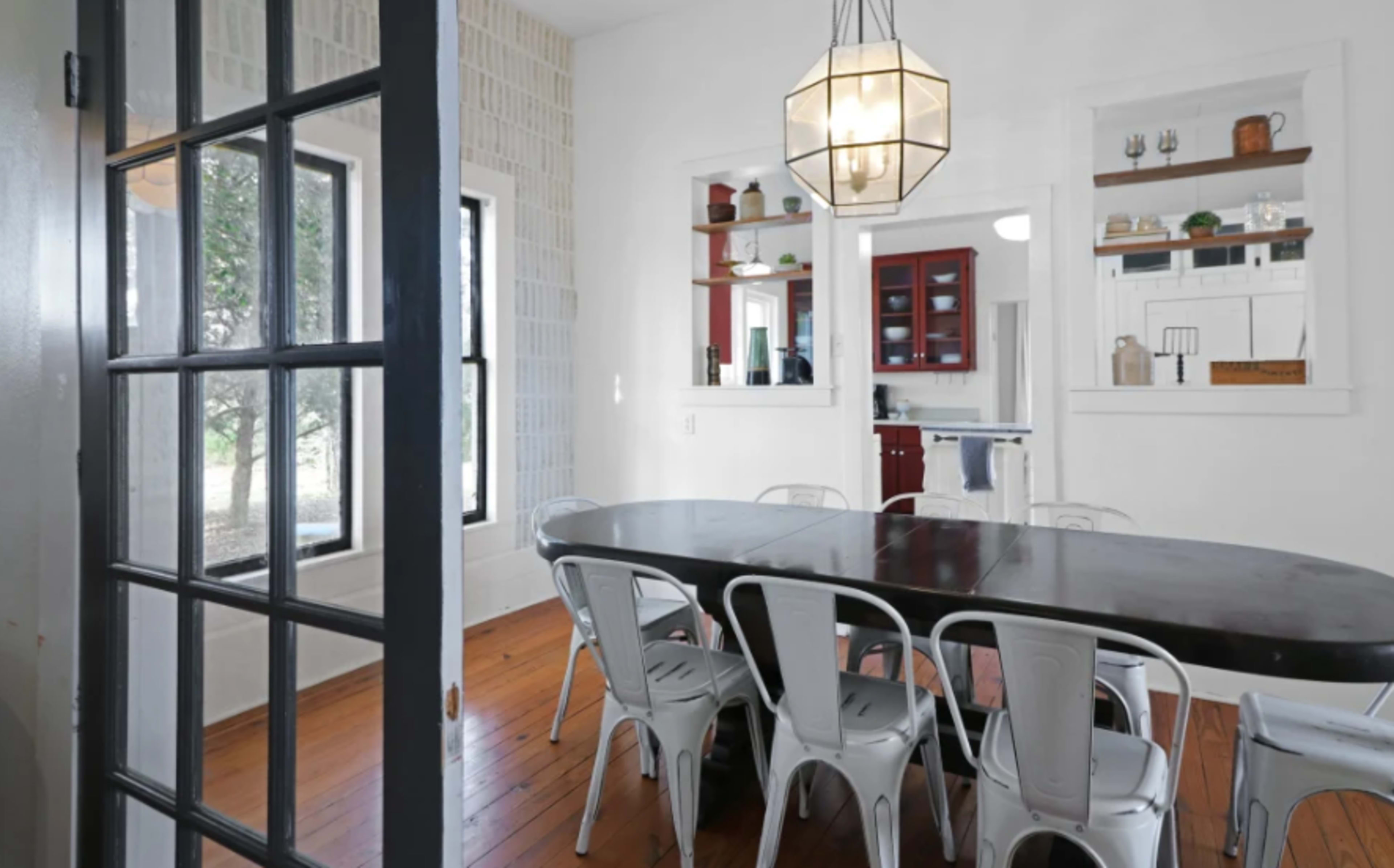 A dining area with a large table surrounded by metal chairs, adjacent to a kitchen and featuring large windows.