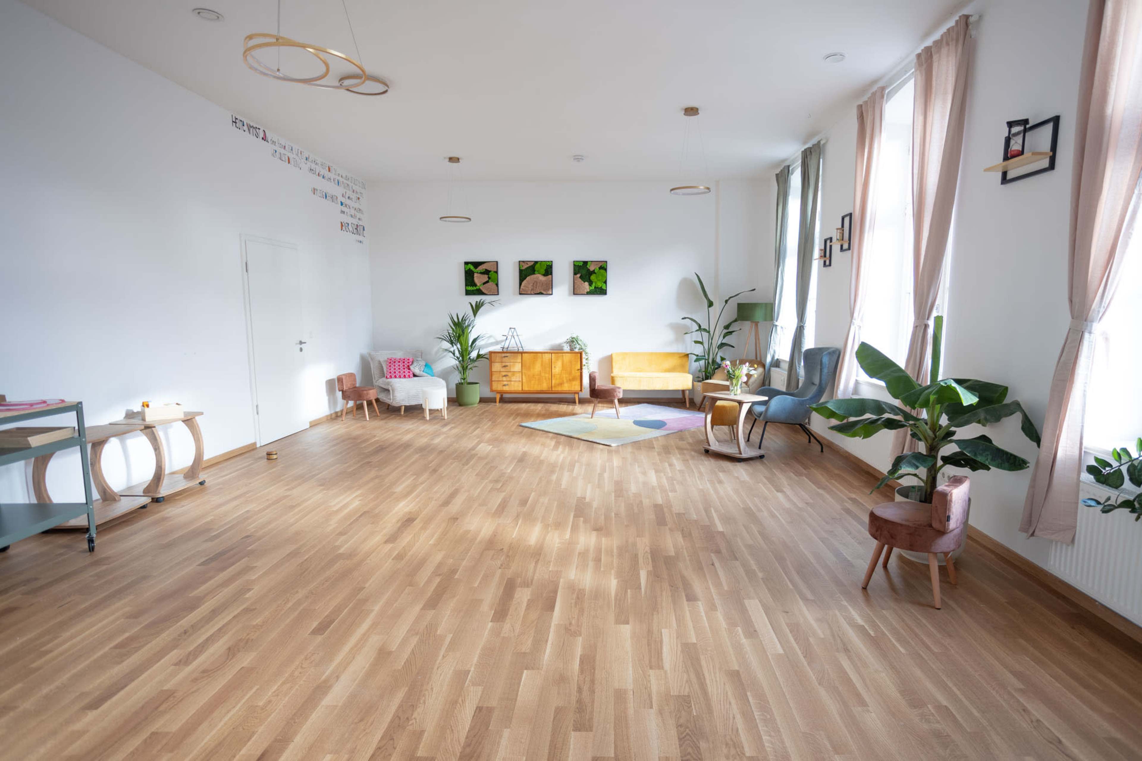 The image shows a spacious, well-lit room with wooden flooring, modern furniture, and potted plants arranged thoughtfully throughout.