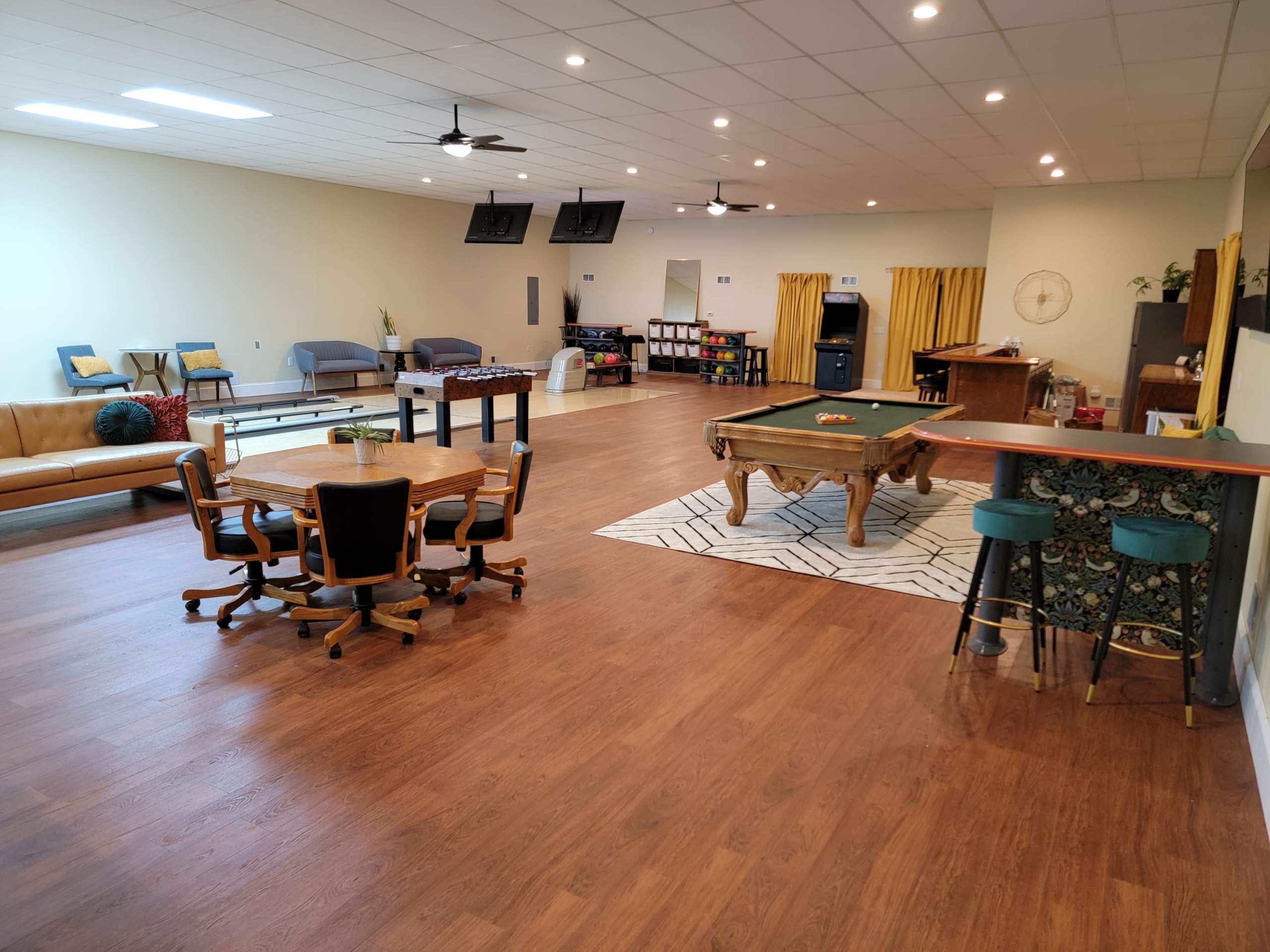 A spacious room with a wooden floor, featuring a pool table, seating areas, and a small kitchen bar.