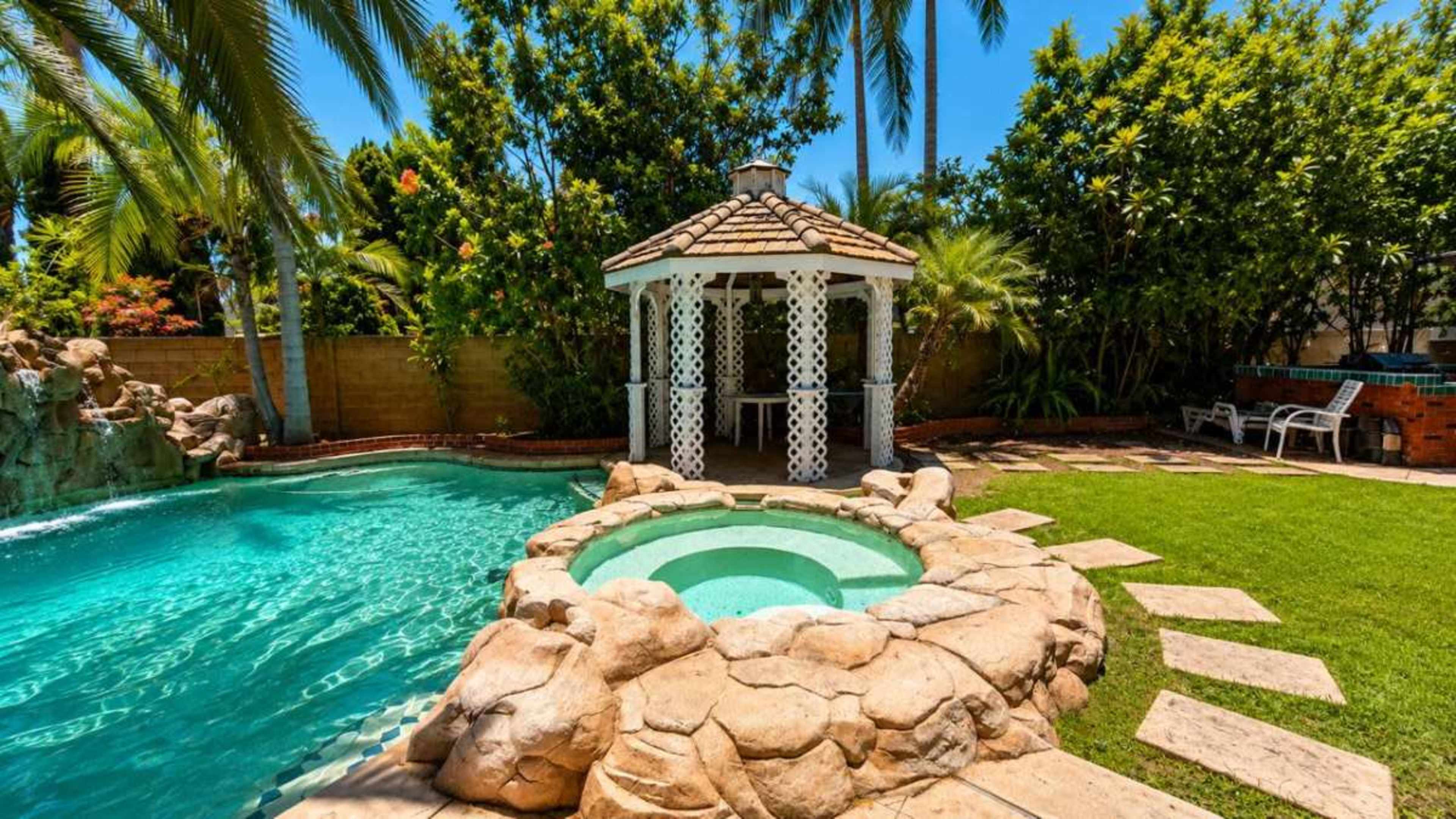 The image shows a tropical pool area featuring a hot tub surrounded by rocks, a gazebo, and lush greenery.