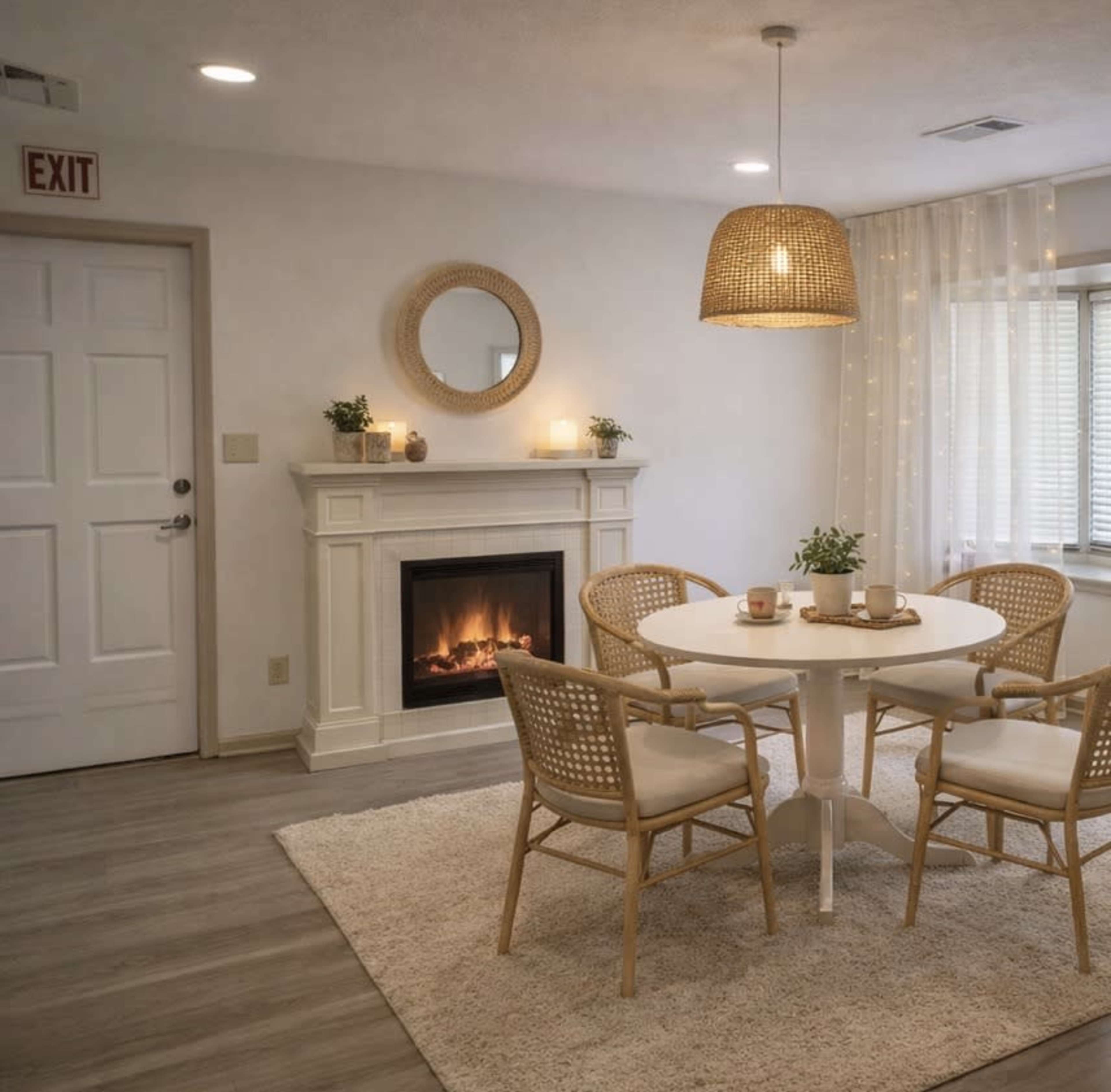 A cozy dining area features a round table with three rattan chairs, positioned near a fireplace, with soft lighting and decorative elements on the walls.