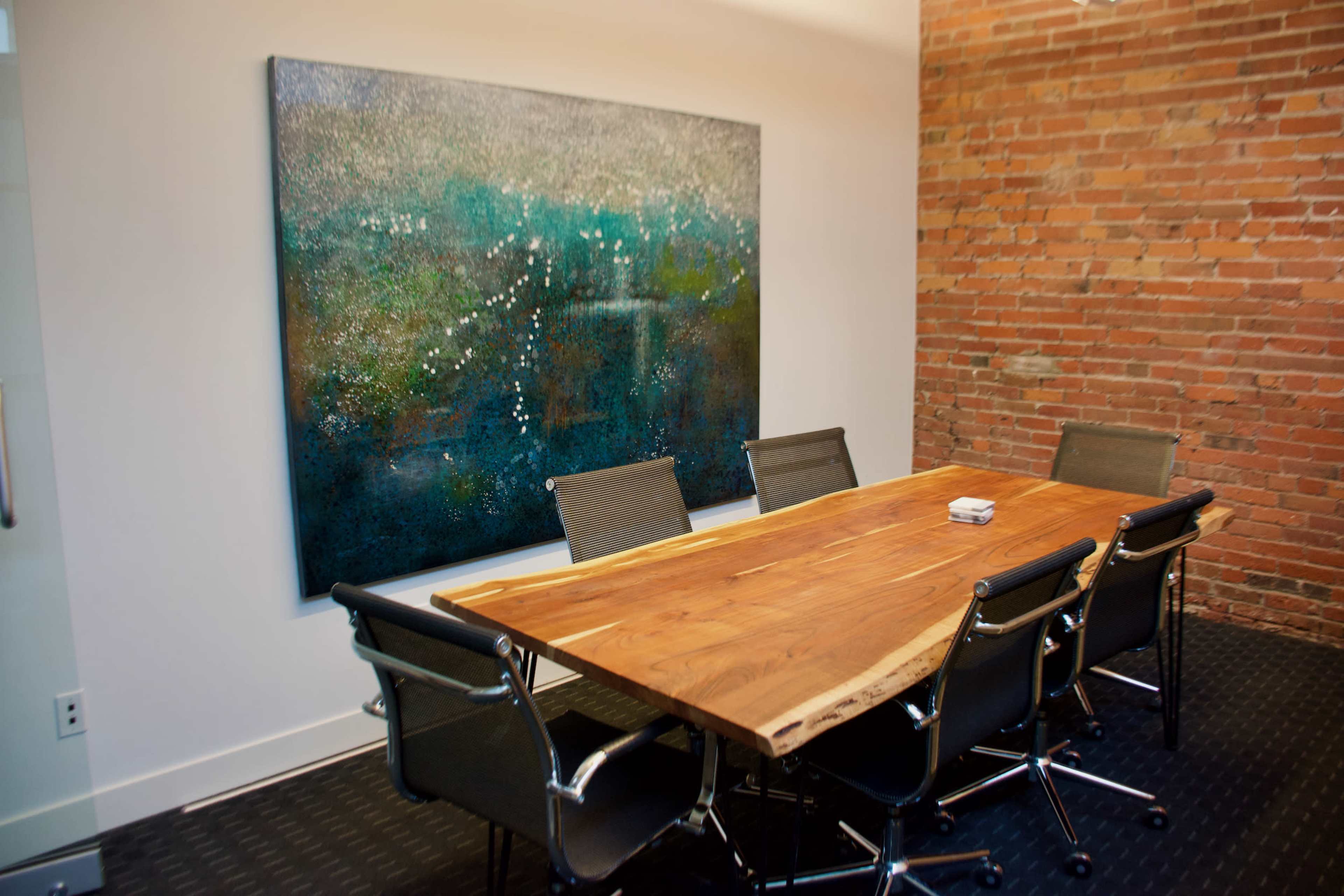 A modern conference room features a large wooden table surrounded by black mesh chairs and a colorful abstract painting on the brick wall.