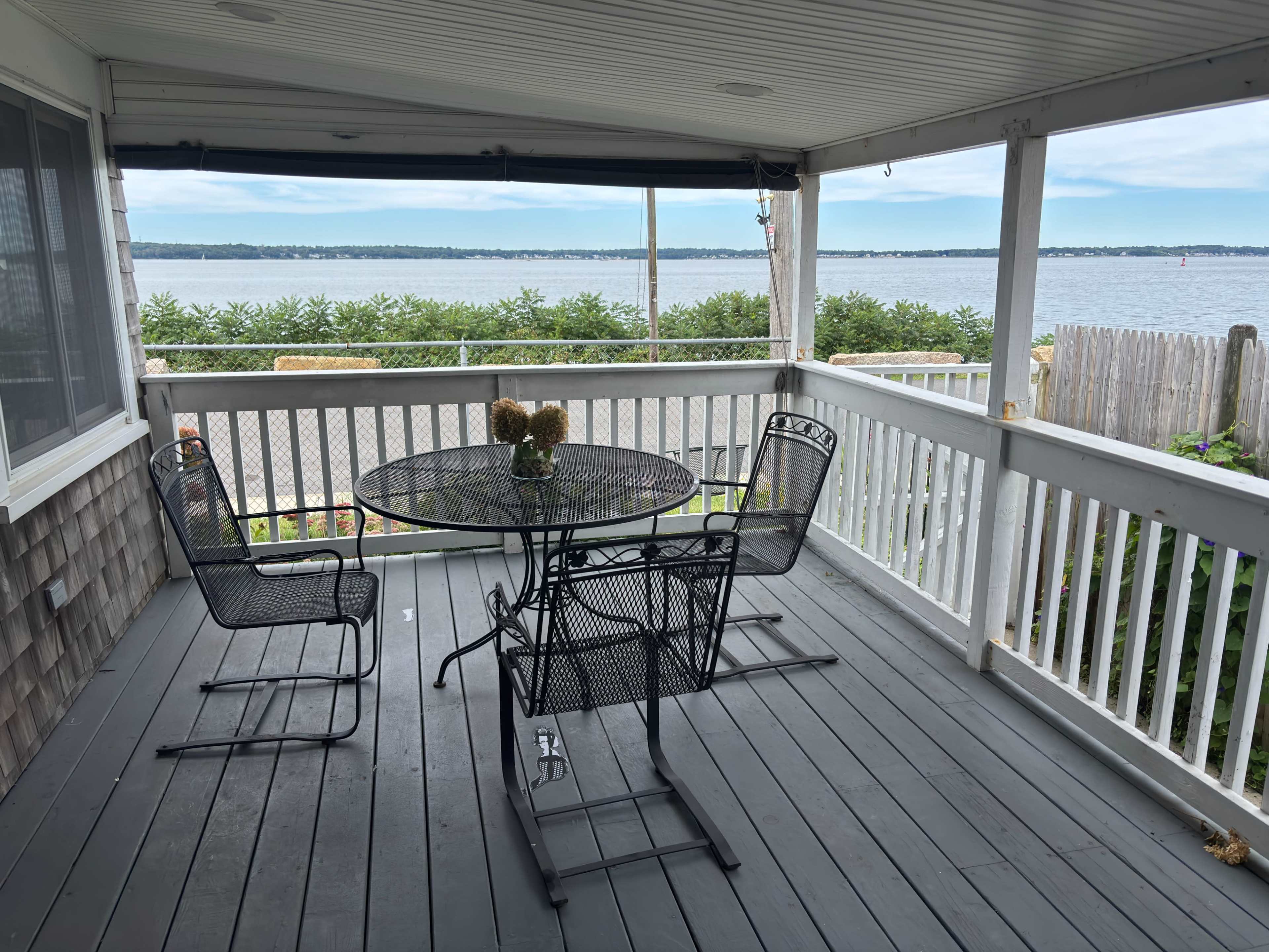 The image shows a porch with a round table and four chairs overlooking a body of water and greenery.