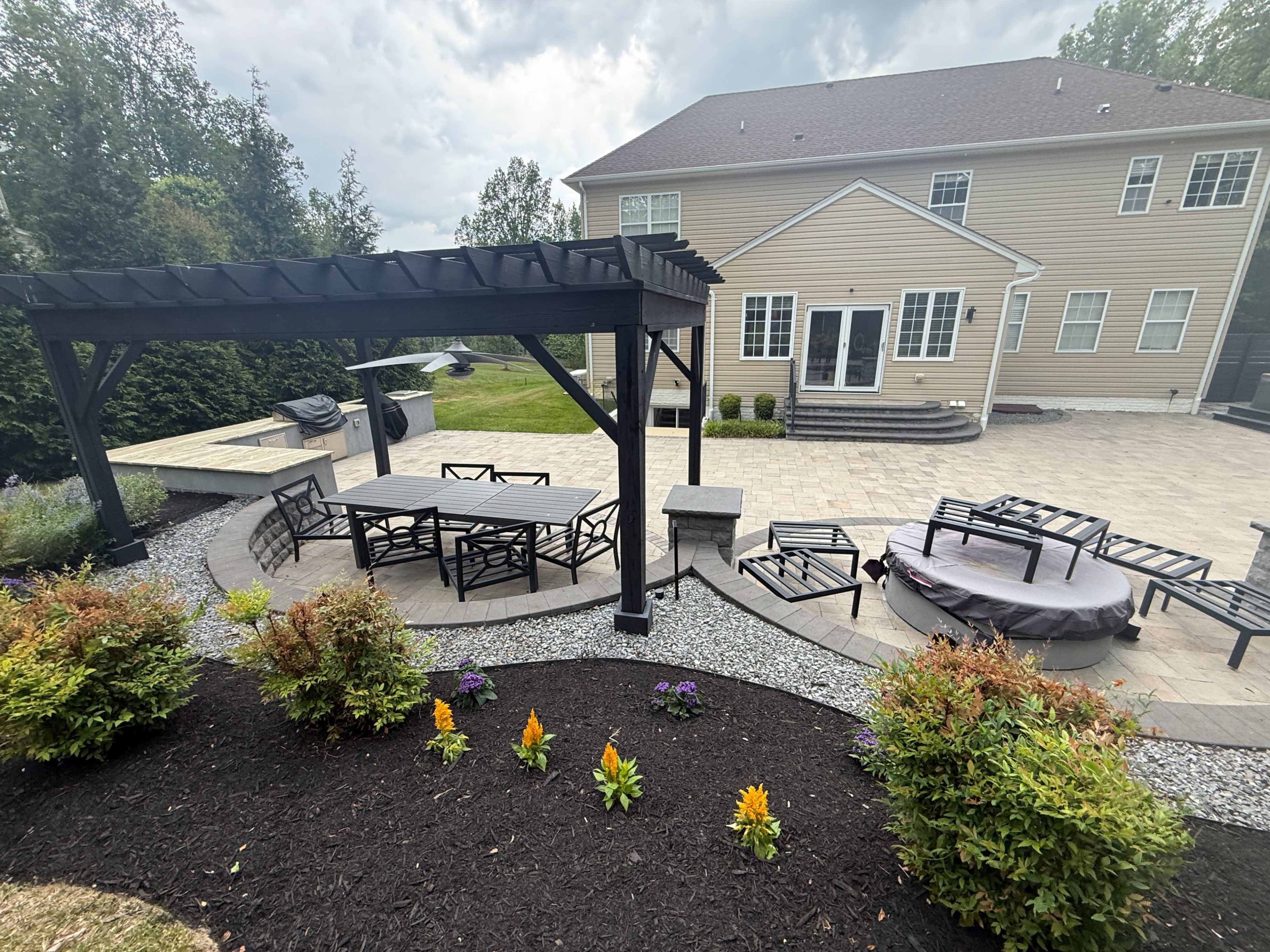 The image shows a patio area with a dining table and chairs under a pergola, surrounded by landscaped gardens and a large house in the background.