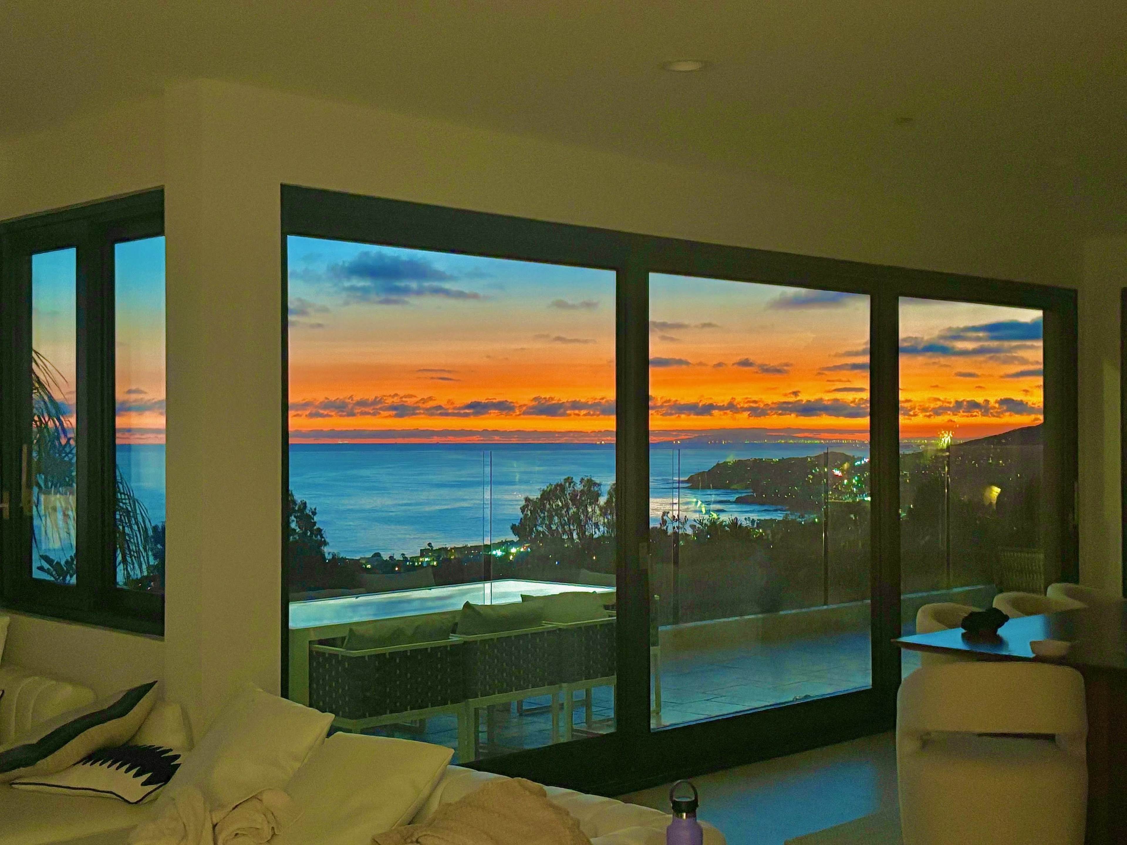 A large glass window frames a colorful sunset over the ocean, with silhouettes of nearby hills and lights in the distance.