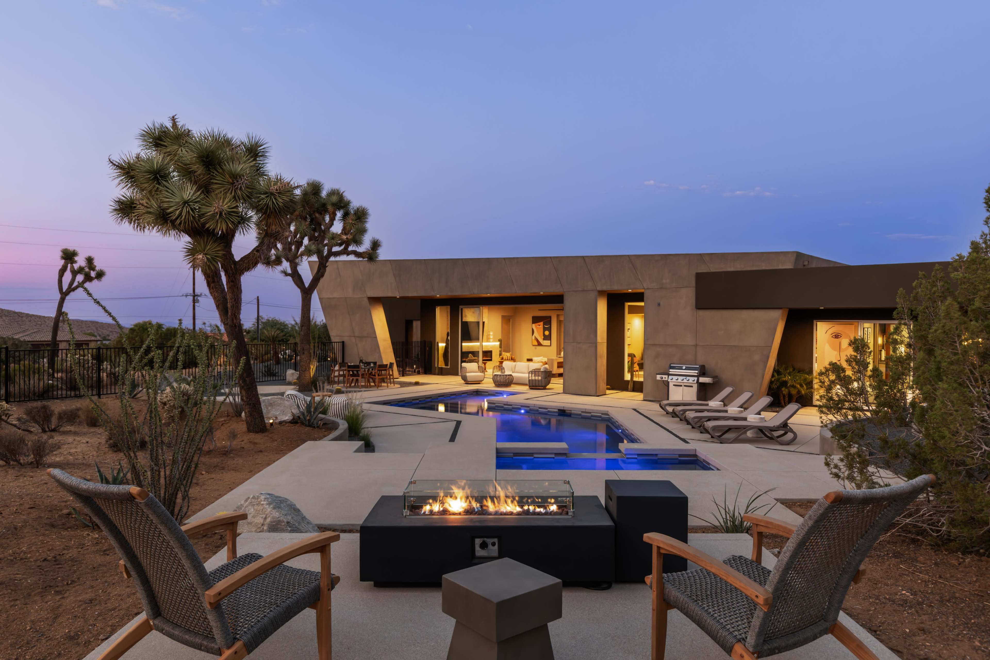 A modern desert home features an outdoor seating area with a fire pit and swimming pool surrounded by Joshua trees and a clear evening sky.