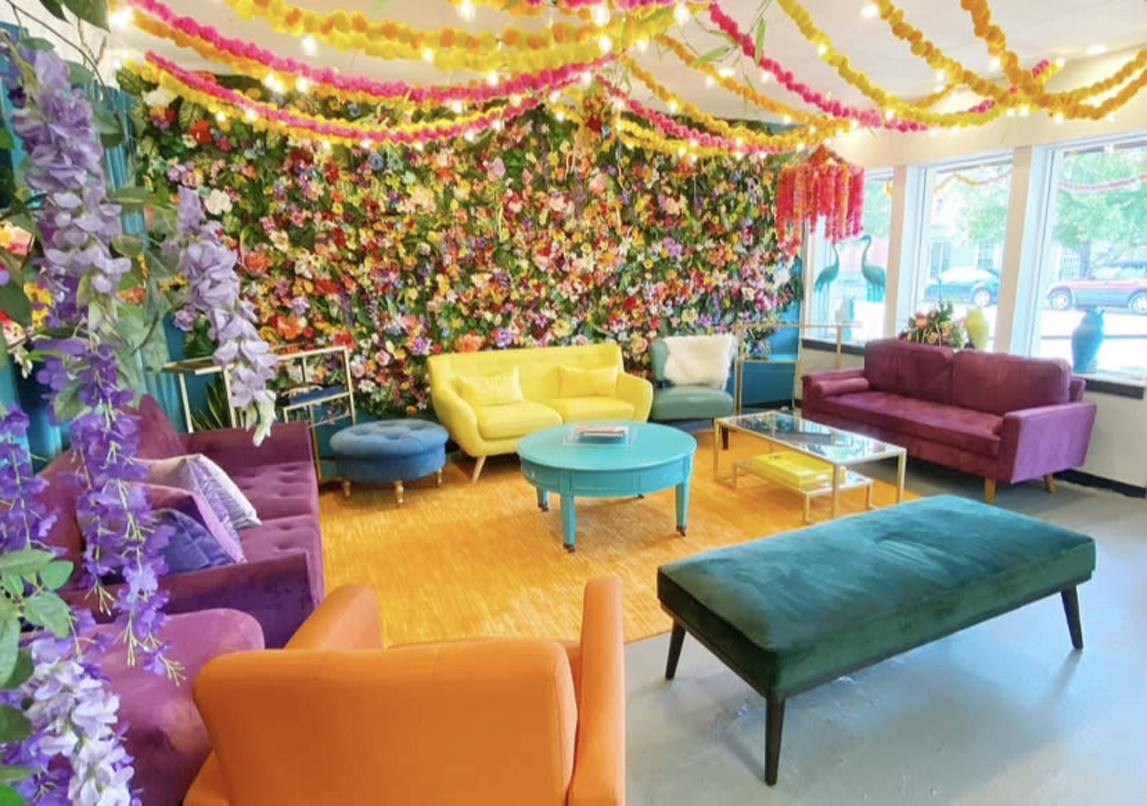 The image shows a vibrant lounge area decorated with a floral wall and various colorful seating options, including sofas and ottomans.