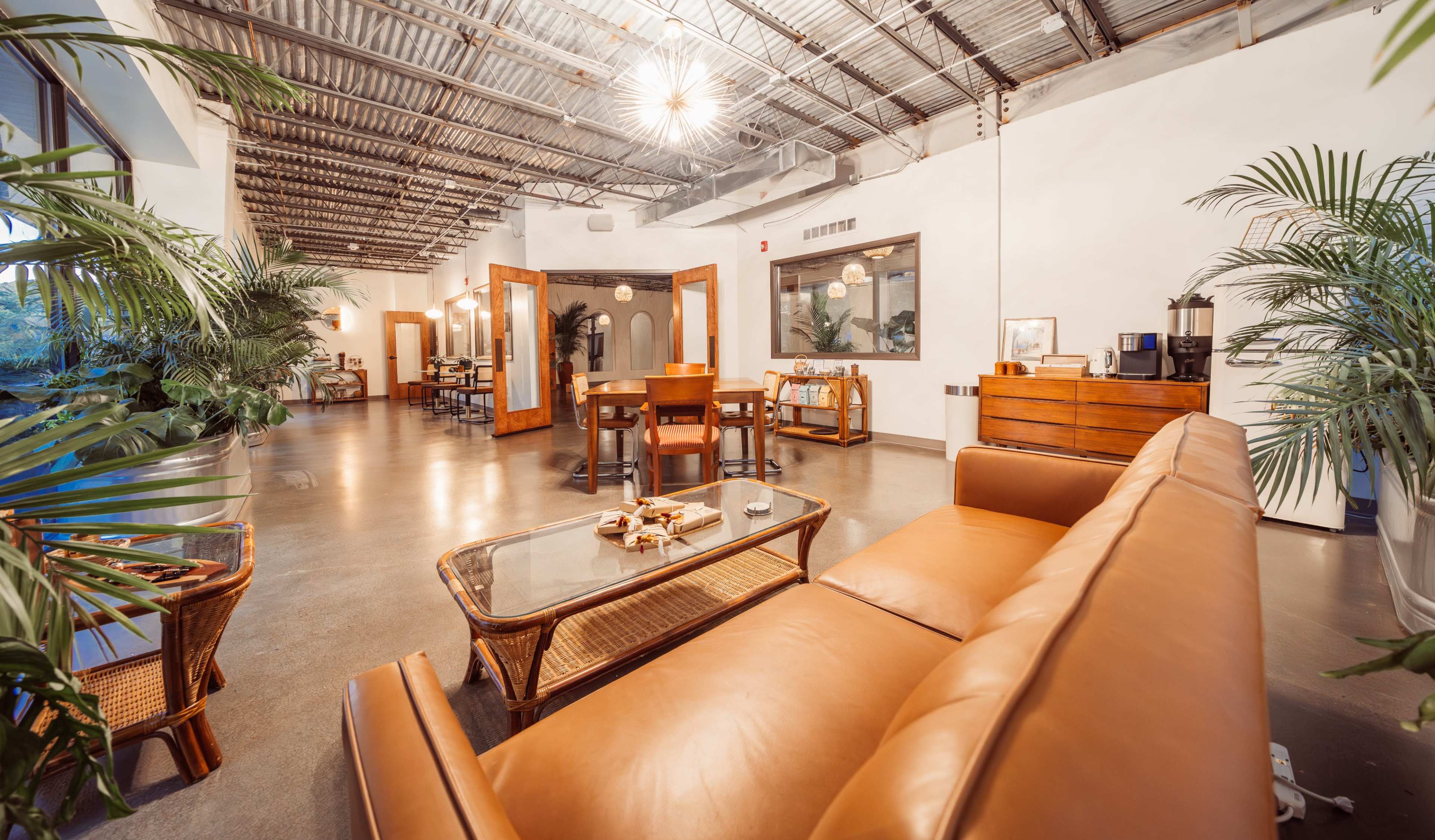 The image shows a modern, spacious lounge area featuring a brown leather sofa, wooden tables, and tropical plants, with warm lighting fixtures overhead.