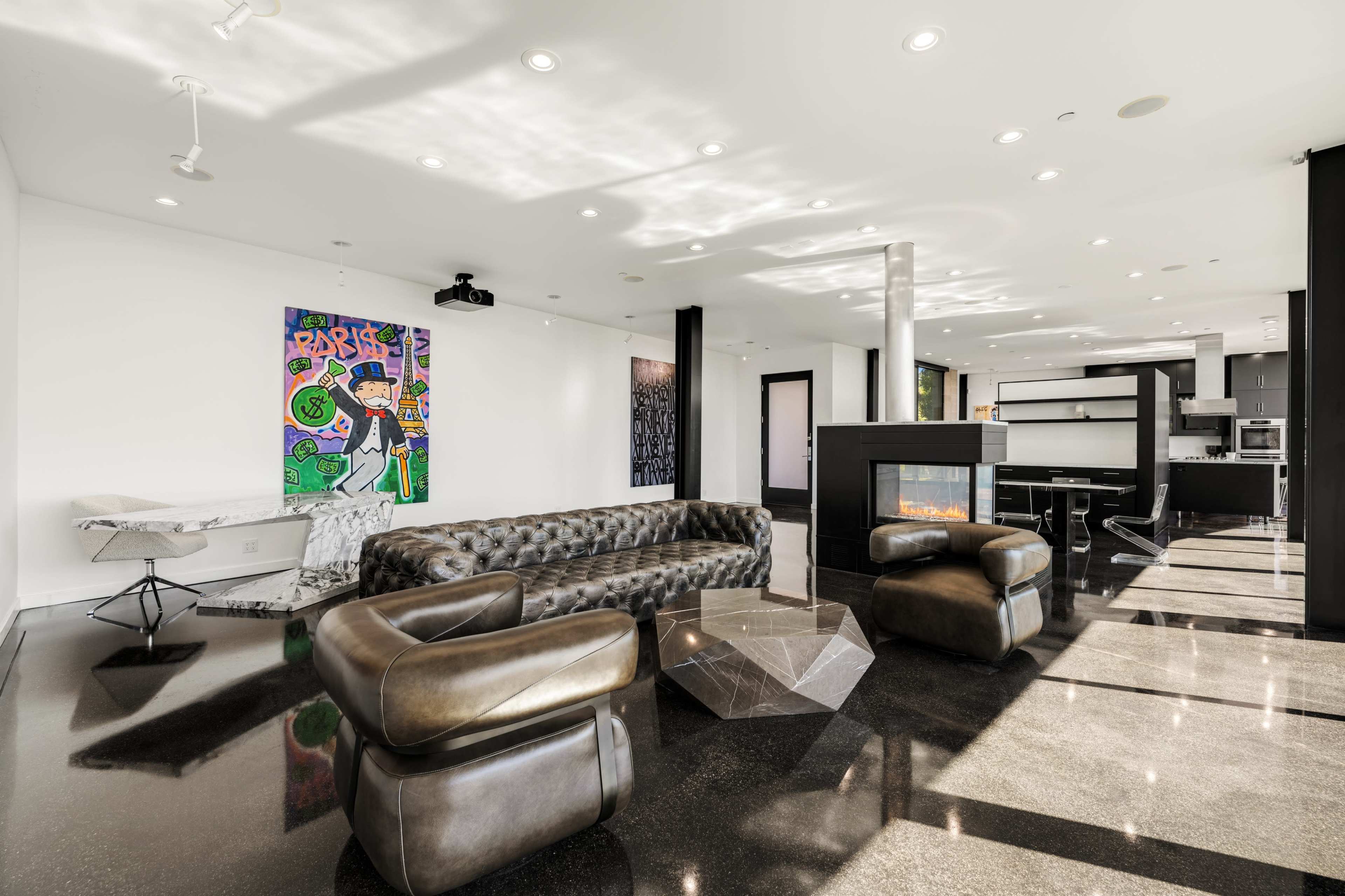 The image shows a modern living space featuring a large leather sofa, an abstract coffee table, and a fireplace, with artwork displayed on the walls and an open kitchen area visible in the background.