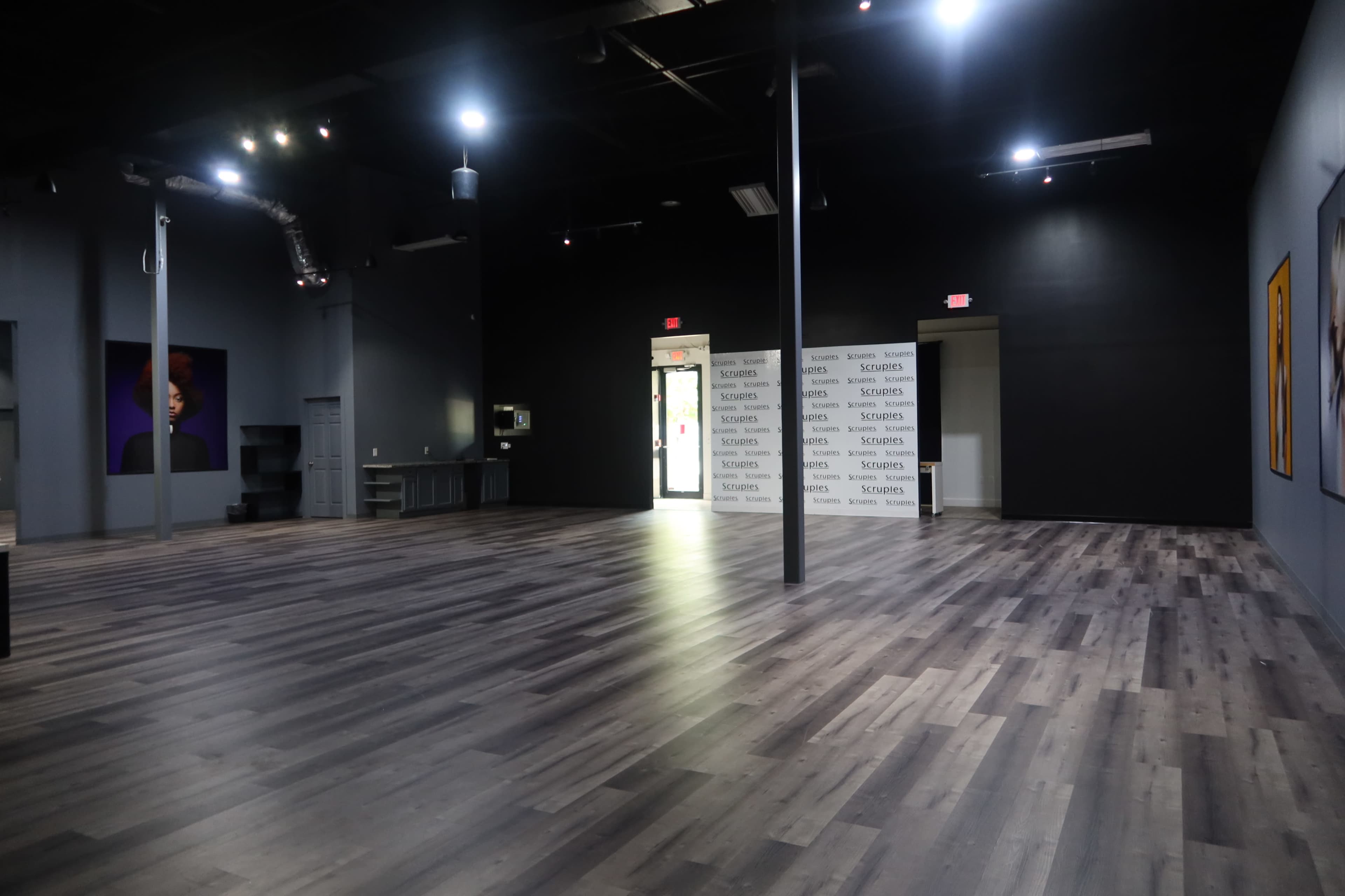 The image shows a spacious, vacant interior of a modern commercial room with dark walls and a wooden floor, featuring a door at one end and a stack of branded materials against a wall.