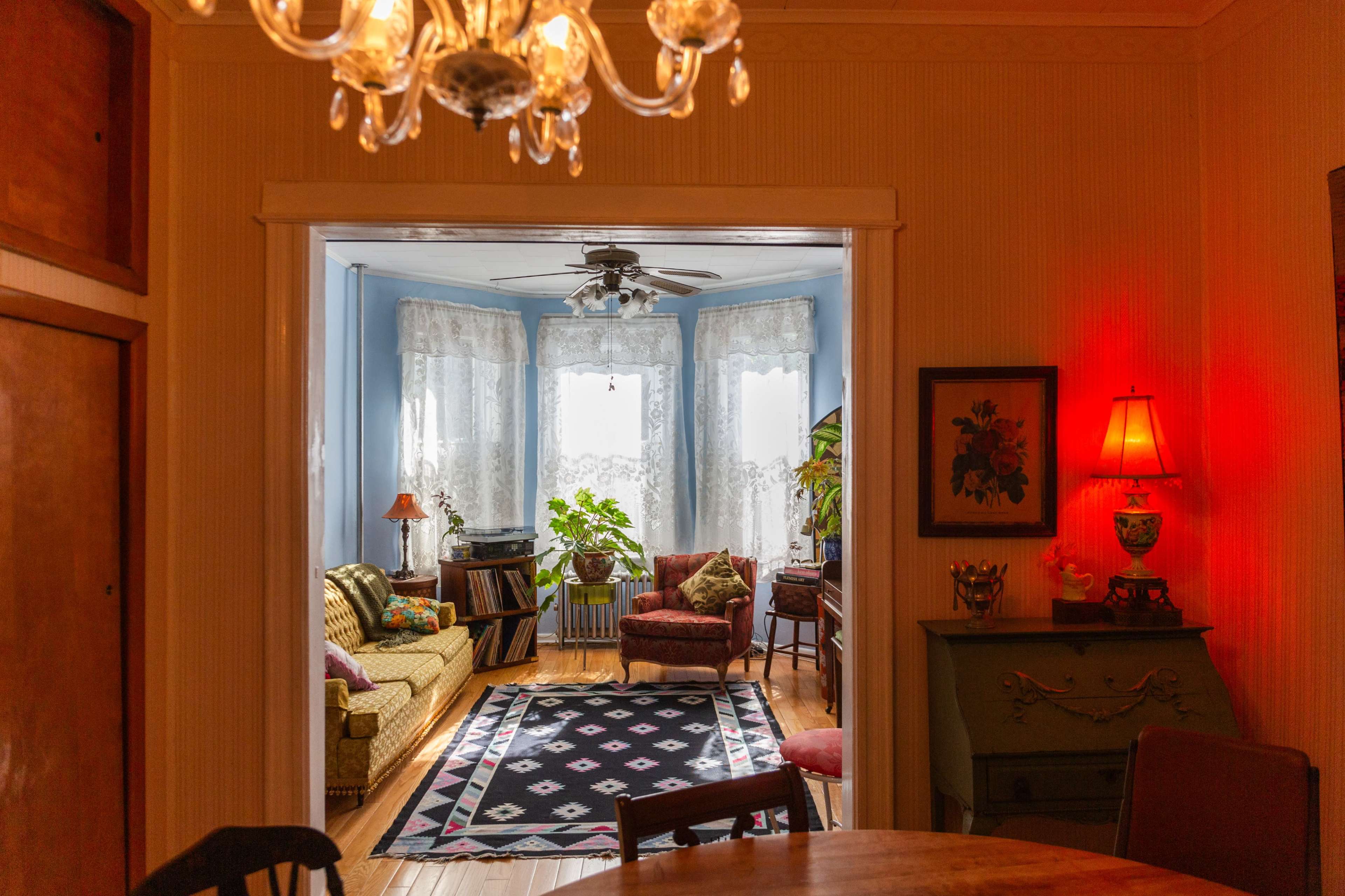 The image shows a cozy room with a living area featuring a patterned rug, comfortable seating, and large window drapes, illuminated by soft lighting.