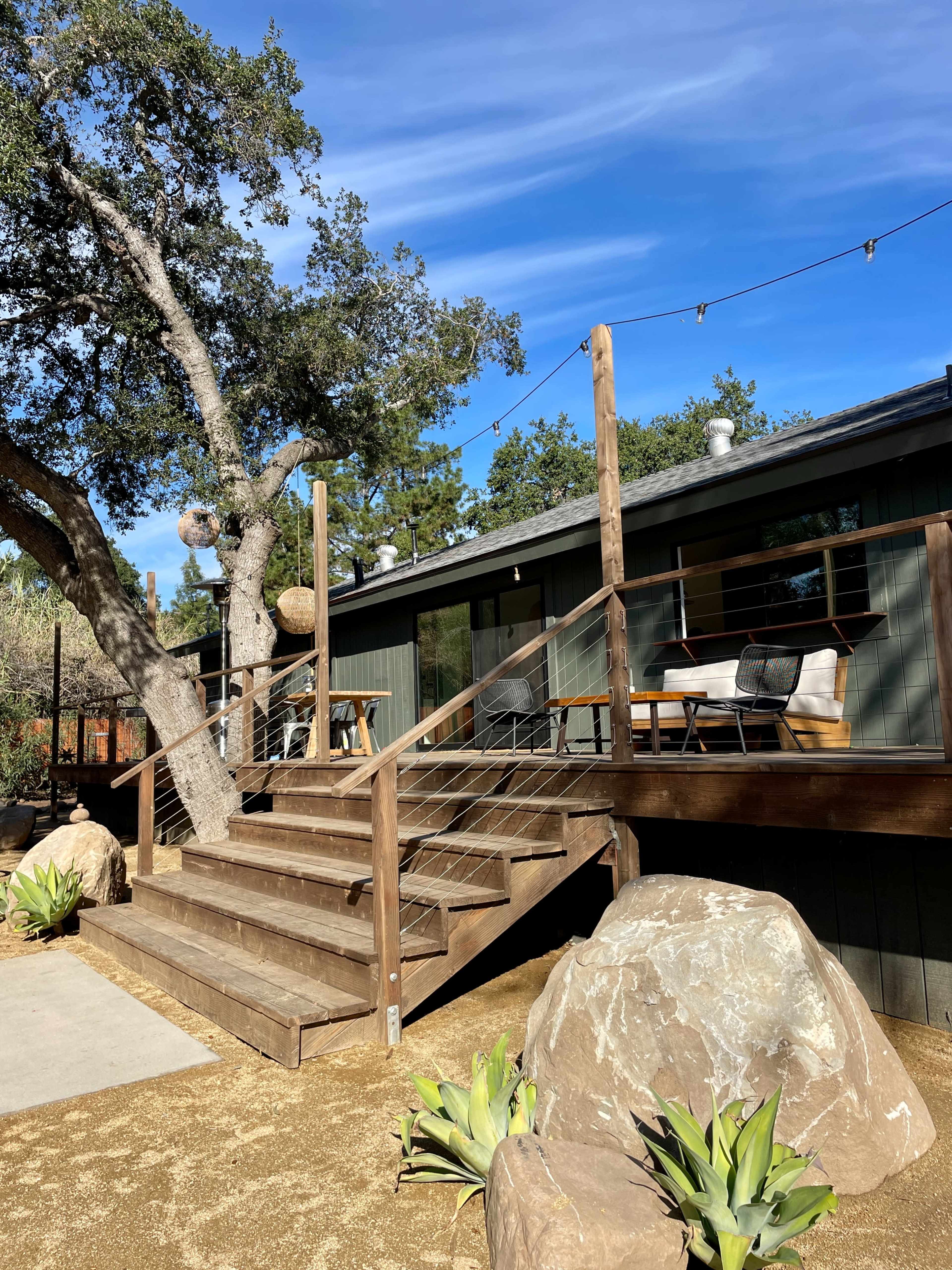 A wooden deck with steps leading up to a green building, surrounded by rocks and desert plants under a blue sky.