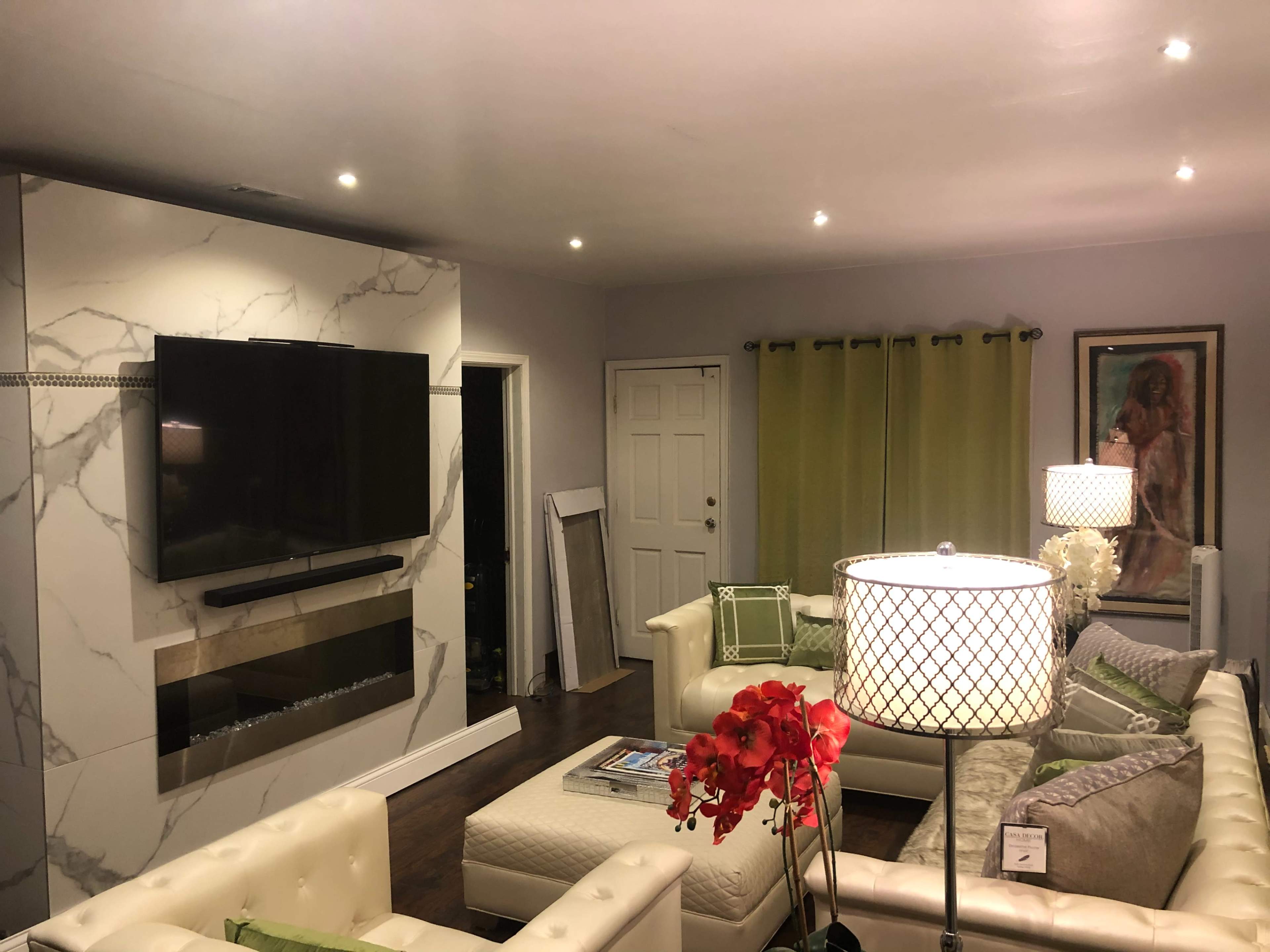 The image shows a modern living room with white sofas, a marble accent wall featuring a flat-screen TV, and decorative lighting.