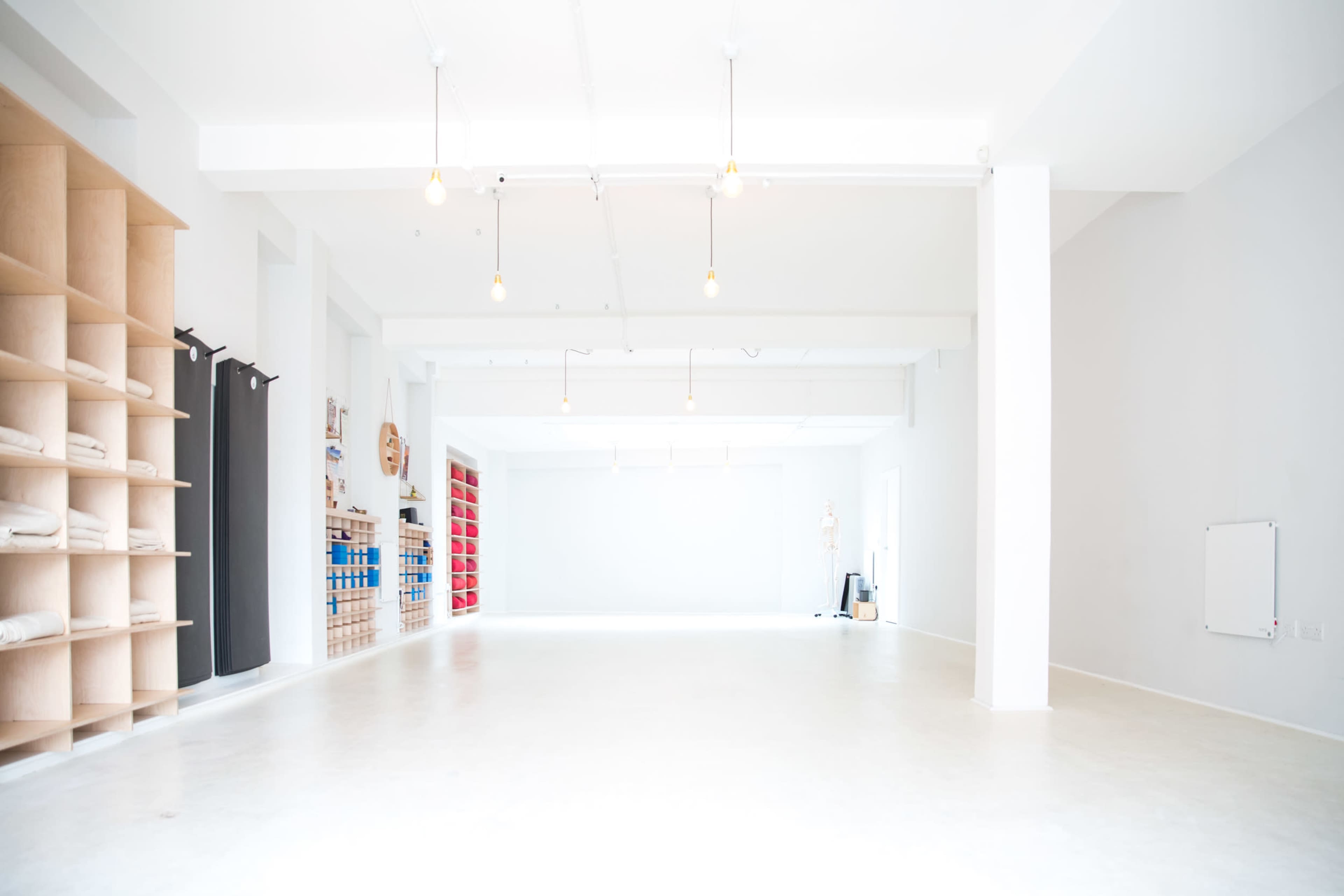 The image shows a spacious, minimalist interior with white walls, wooden shelving on one side, and rows of overhead lights.