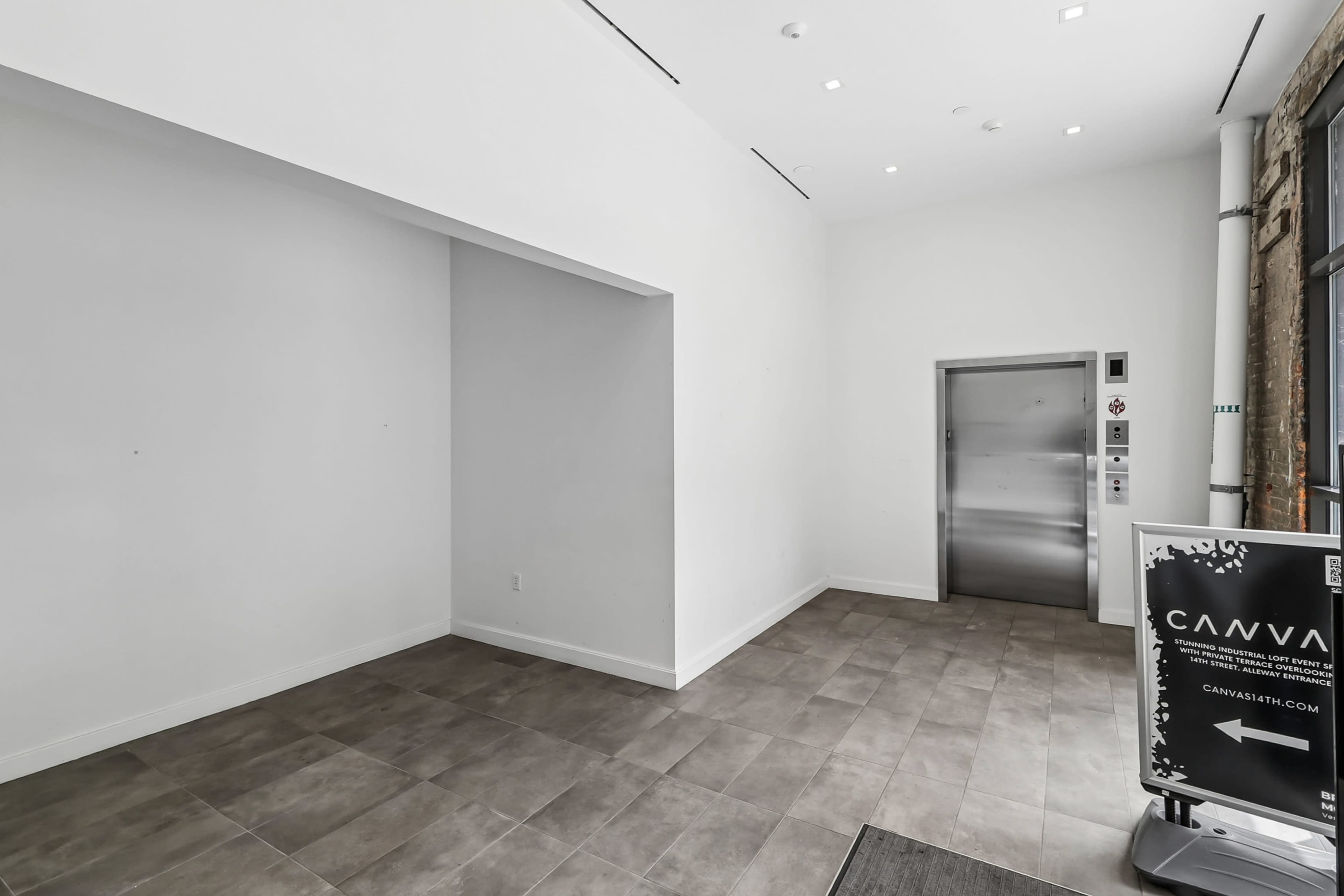 The image shows a spacious, bright lobby area with gray tiled flooring and an elevator to the right, accompanied by a signage stand on the left.