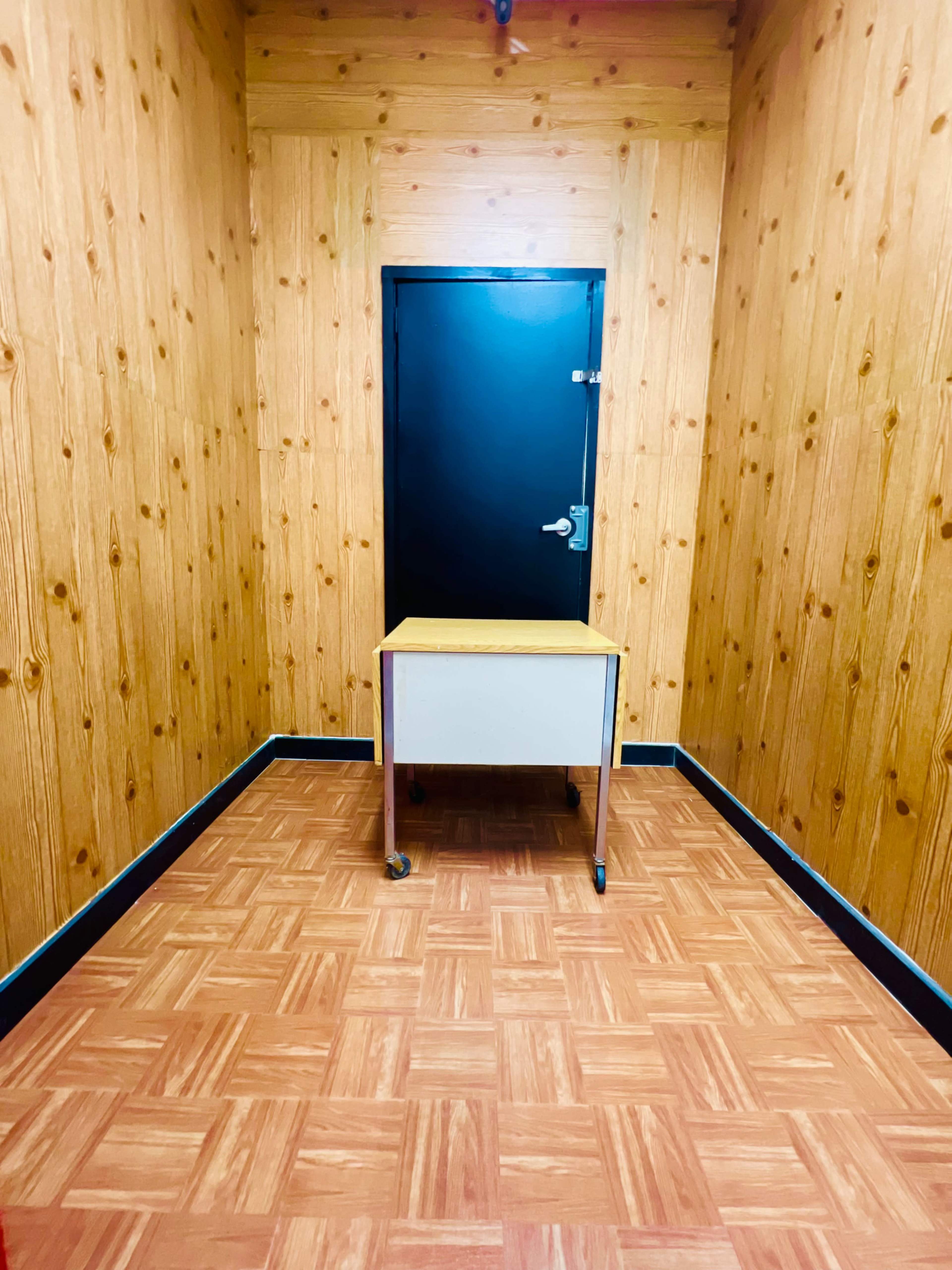 A small room with wooden walls features a single black door and a table placed in the center on tiled flooring.