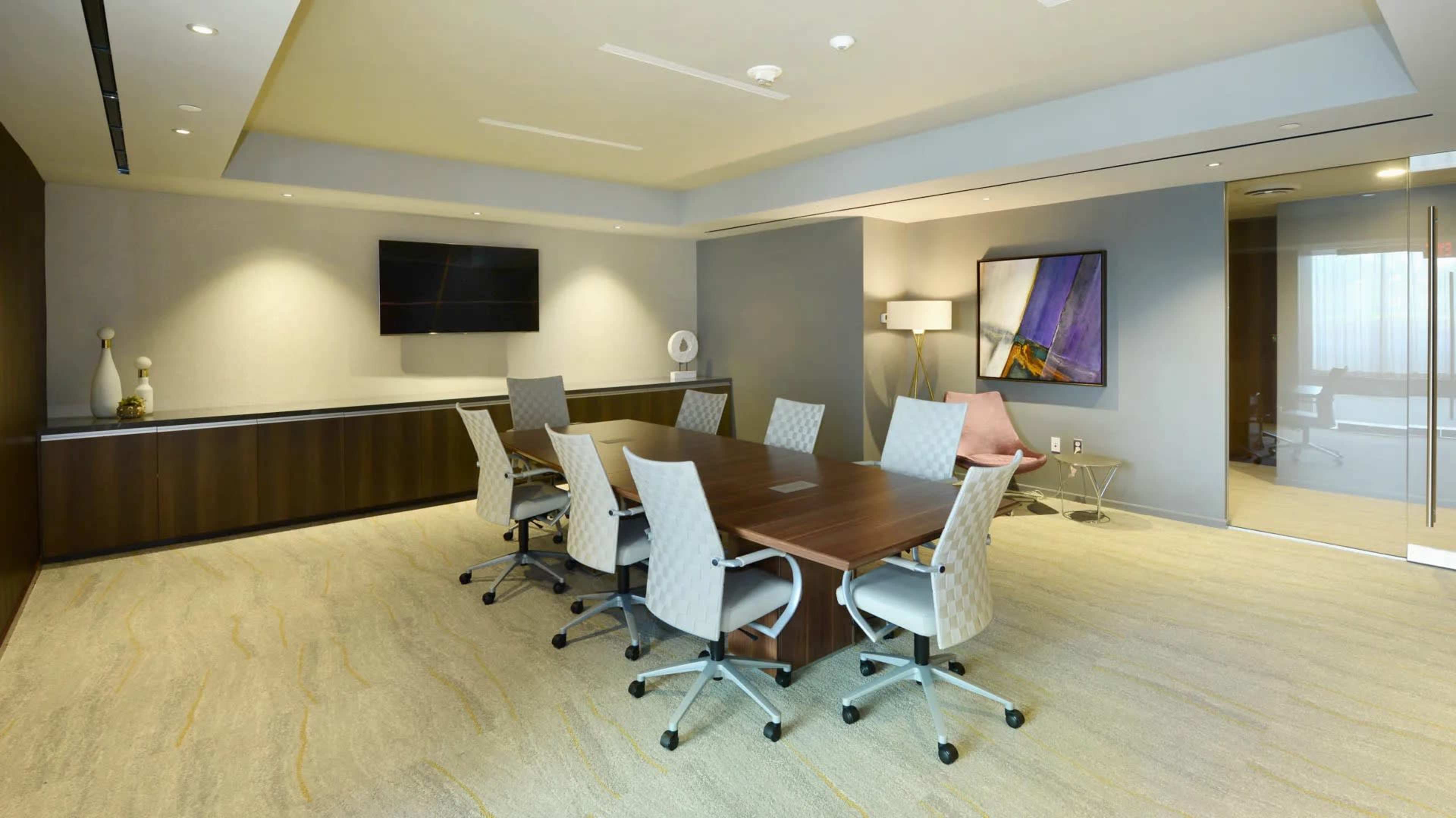 A modern conference room features a large wooden table surrounded by six ergonomic chairs, with a television mounted on the wall and a glass partition leading to another space.
