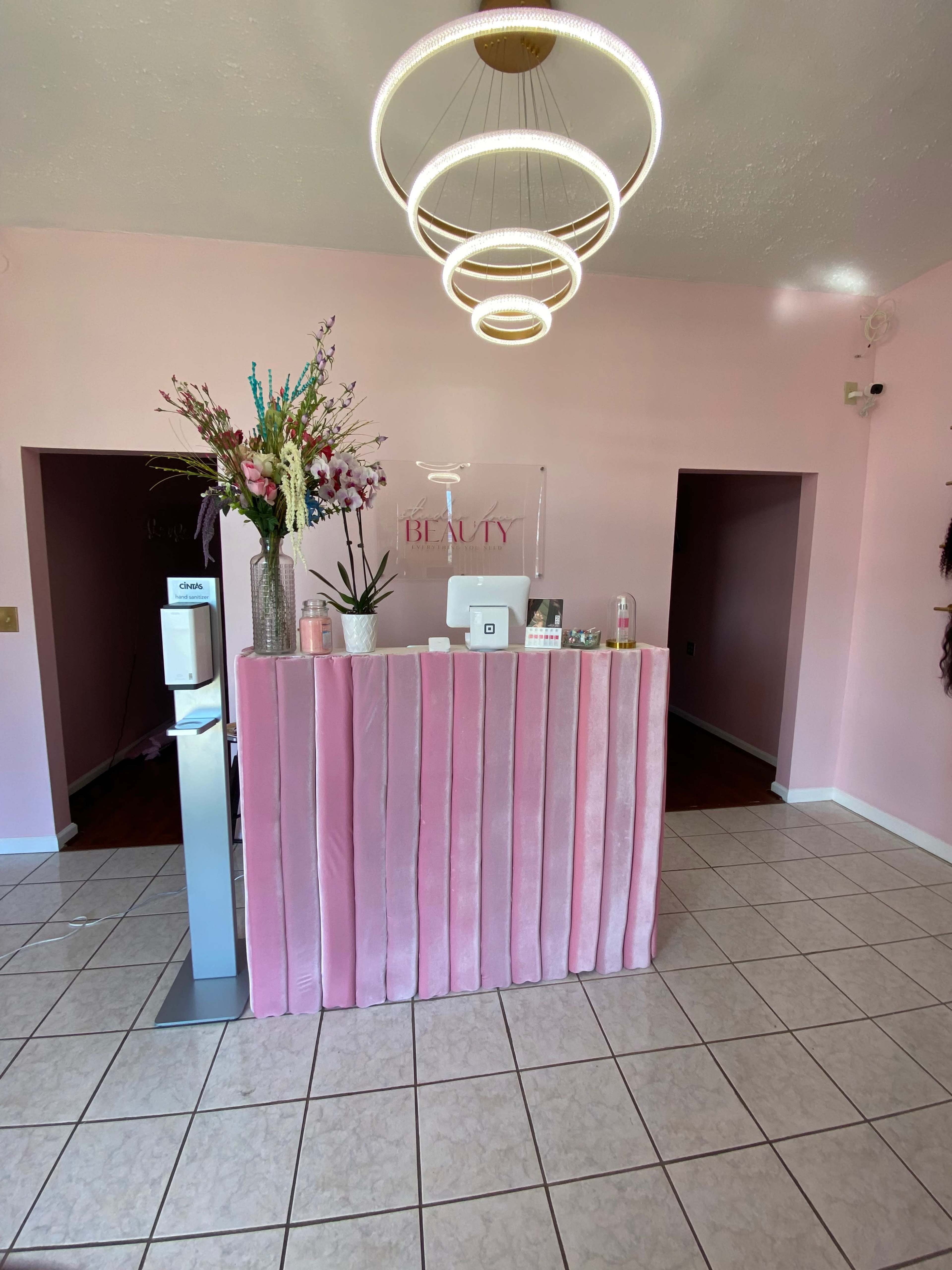 A beauty reception area featuring a pink velvet counter, decorative flowers, and modern light fixtures against a pink wall.