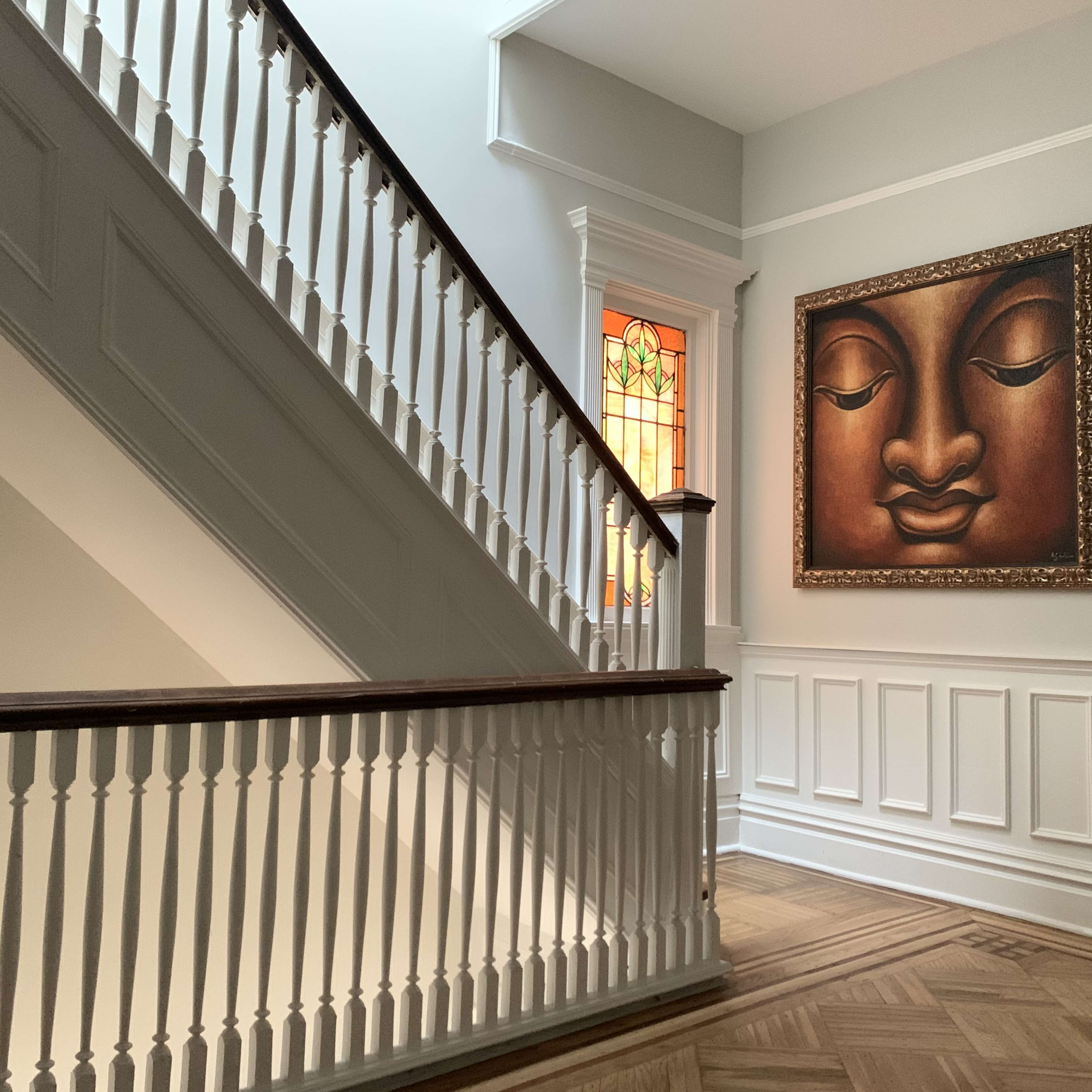 A stairway with a wooden handrail and a large painting of a Buddha face on the wall beside a stained glass window.