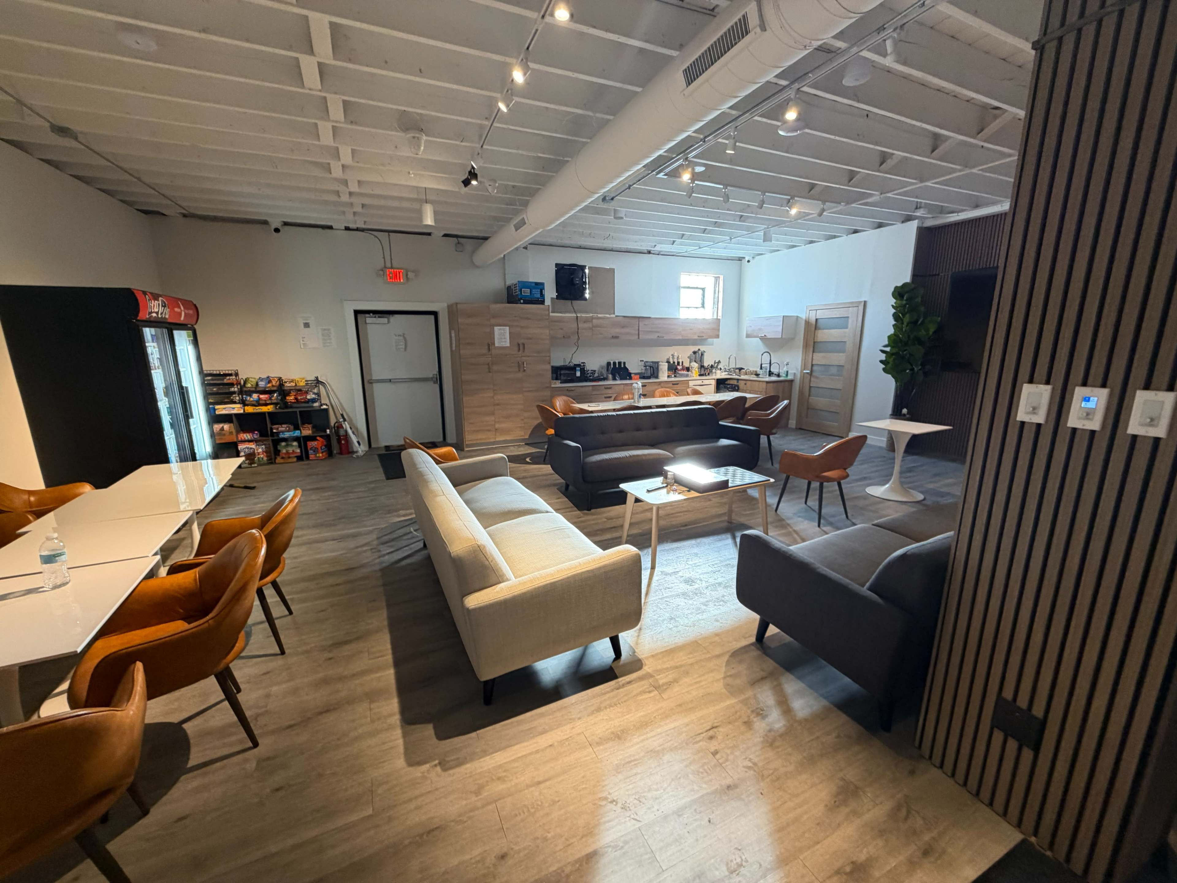 The image shows a modern communal kitchen and lounge area with multiple seating options, a kitchenette, and a refrigerator stocked with snacks.
