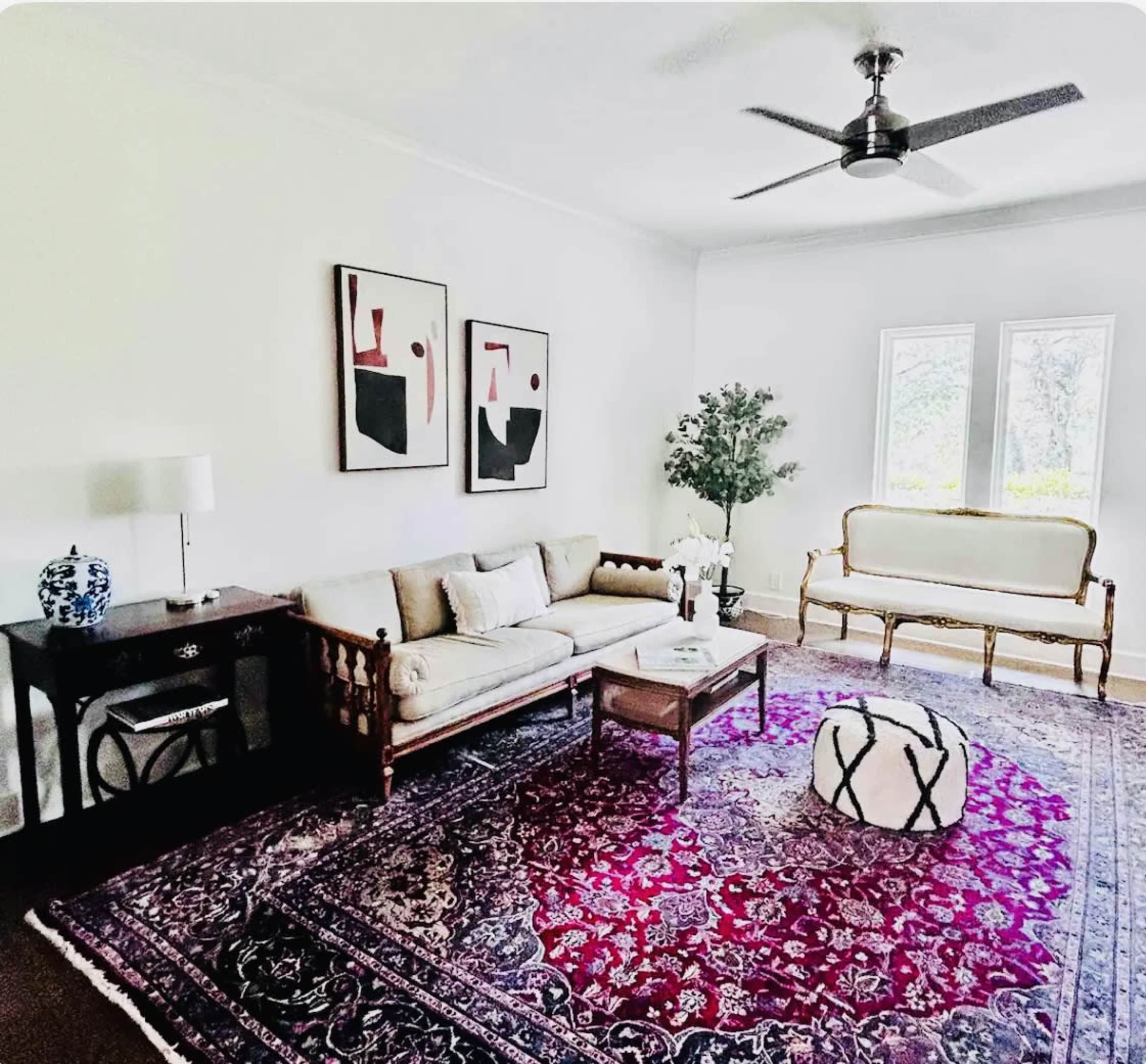 The living room features a beige sofa, a wooden side table, a patterned area rug, and two framed artworks, with a large window allowing natural light to enter.