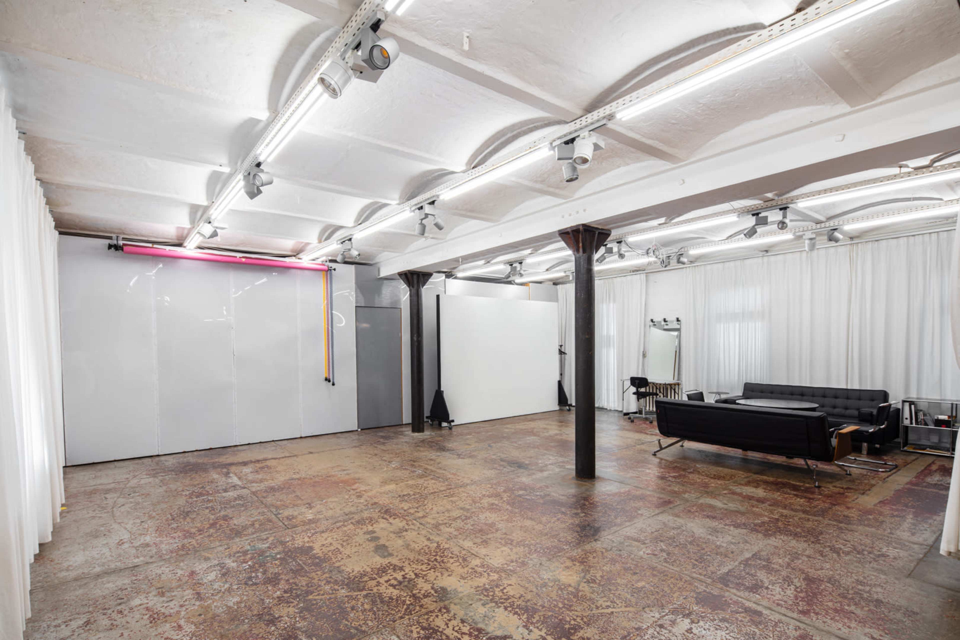 The image shows a spacious studio with polished concrete flooring, white drapes, and minimalist furniture, featuring a black couch and lighting fixtures along the ceiling.