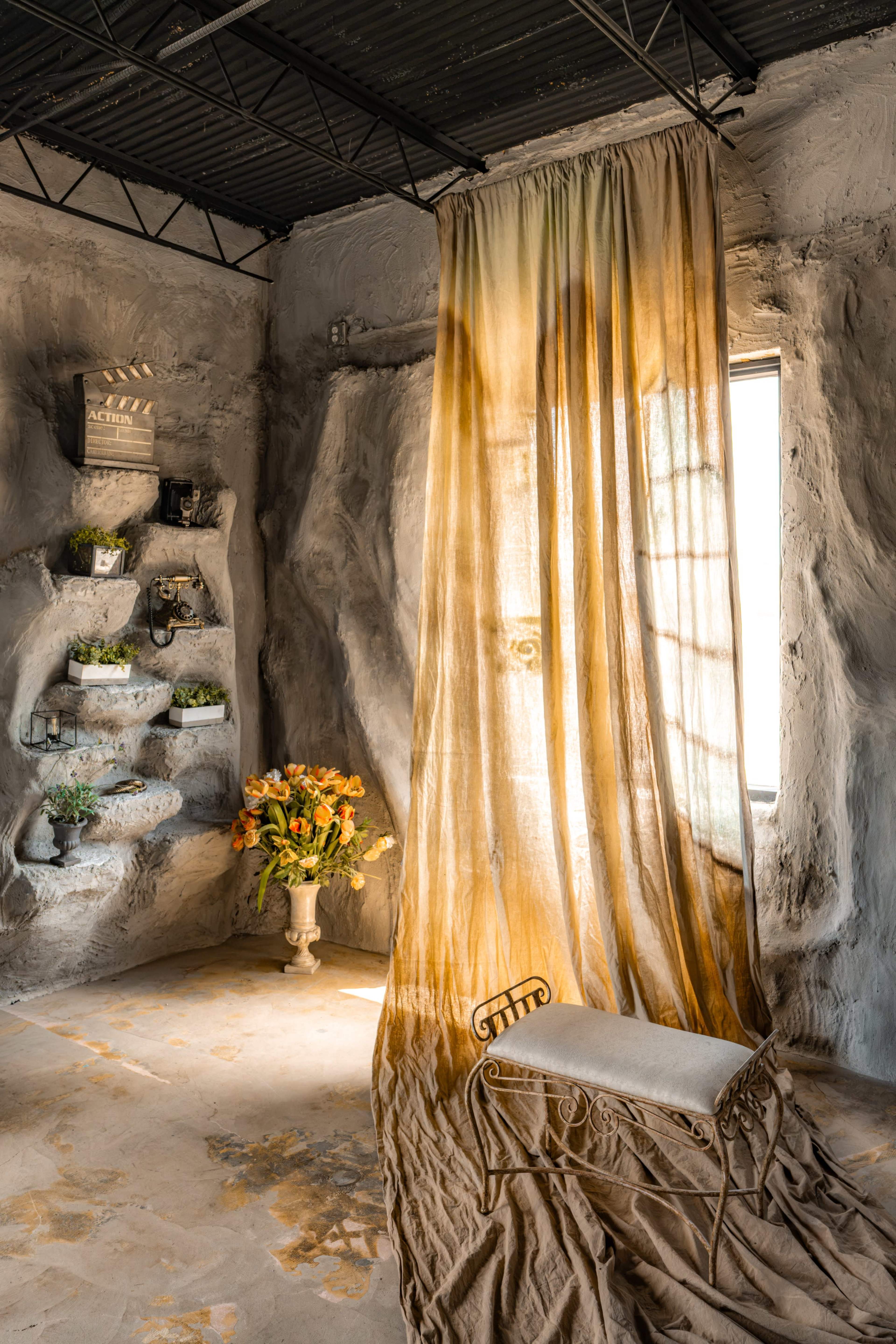 The image shows a room with textured walls, a large curtain draping near a window, a decorative shelf with plants, and a small floral arrangement in a vase.