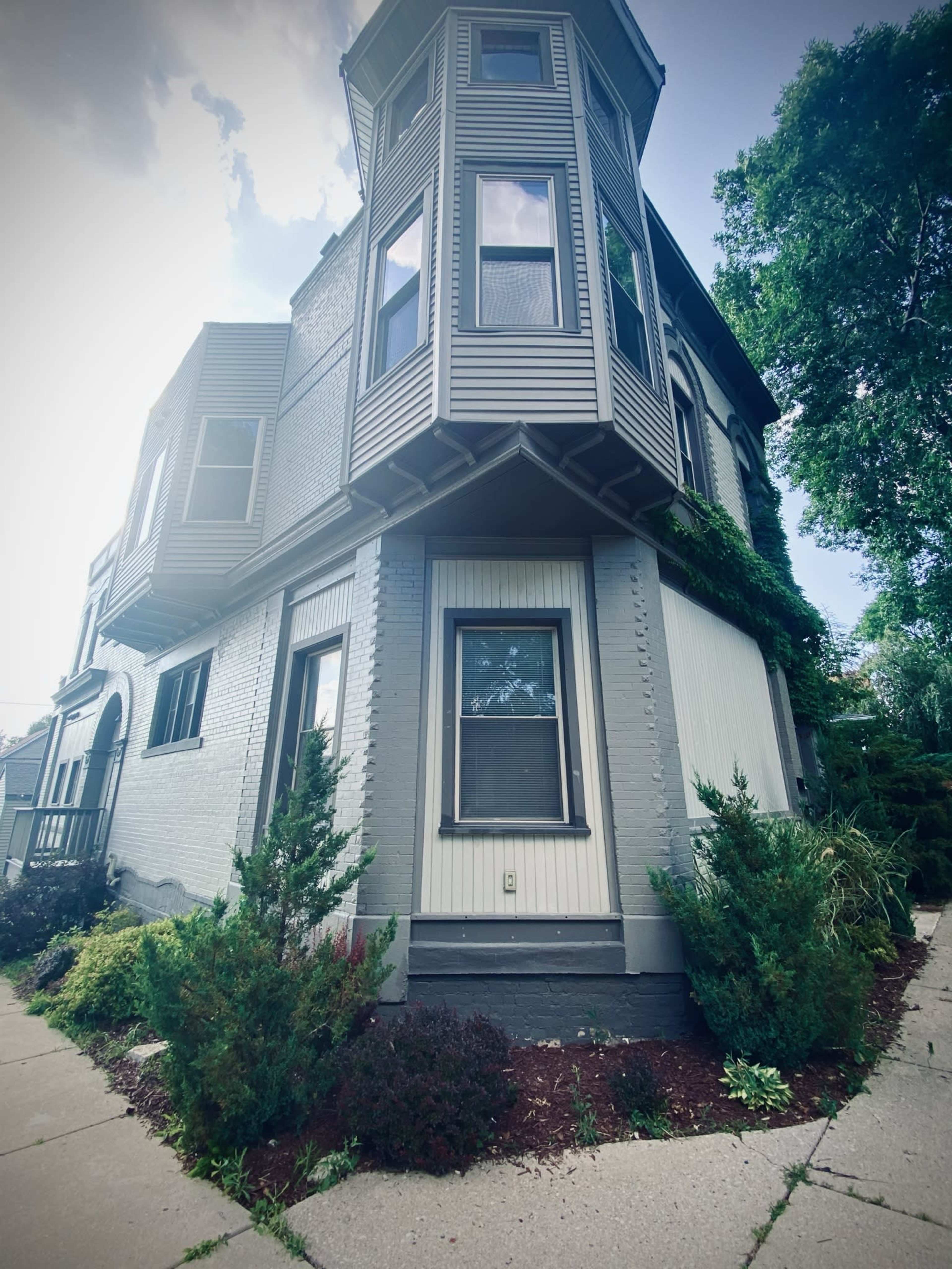 A corner view of a two-story house with a distinct turret, featuring brick and siding elements, alongside a walkway and landscaped shrubs.