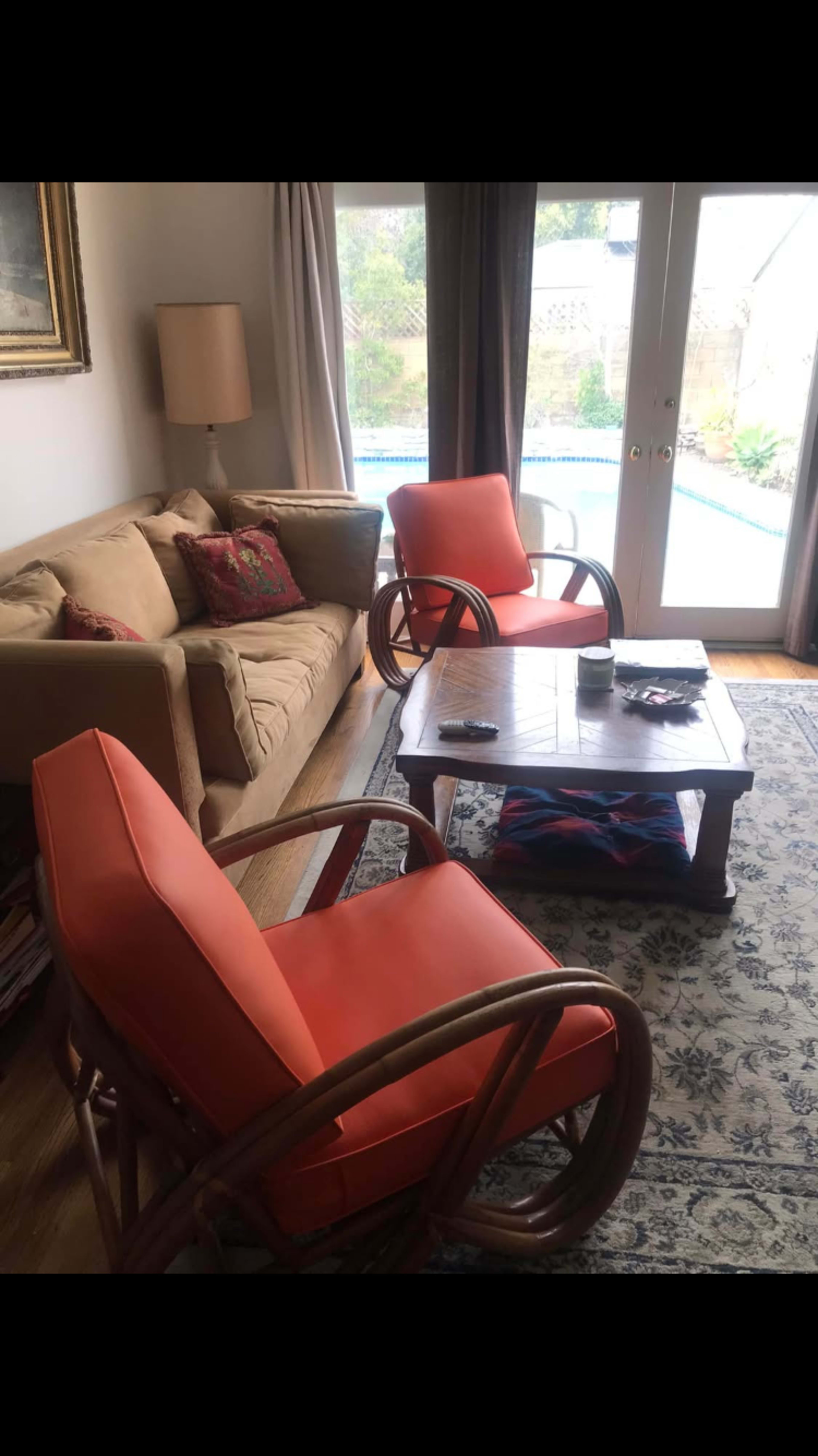 The living area features two orange chairs, a wooden coffee table, a beige couch, and large windows overlooking a pool area.