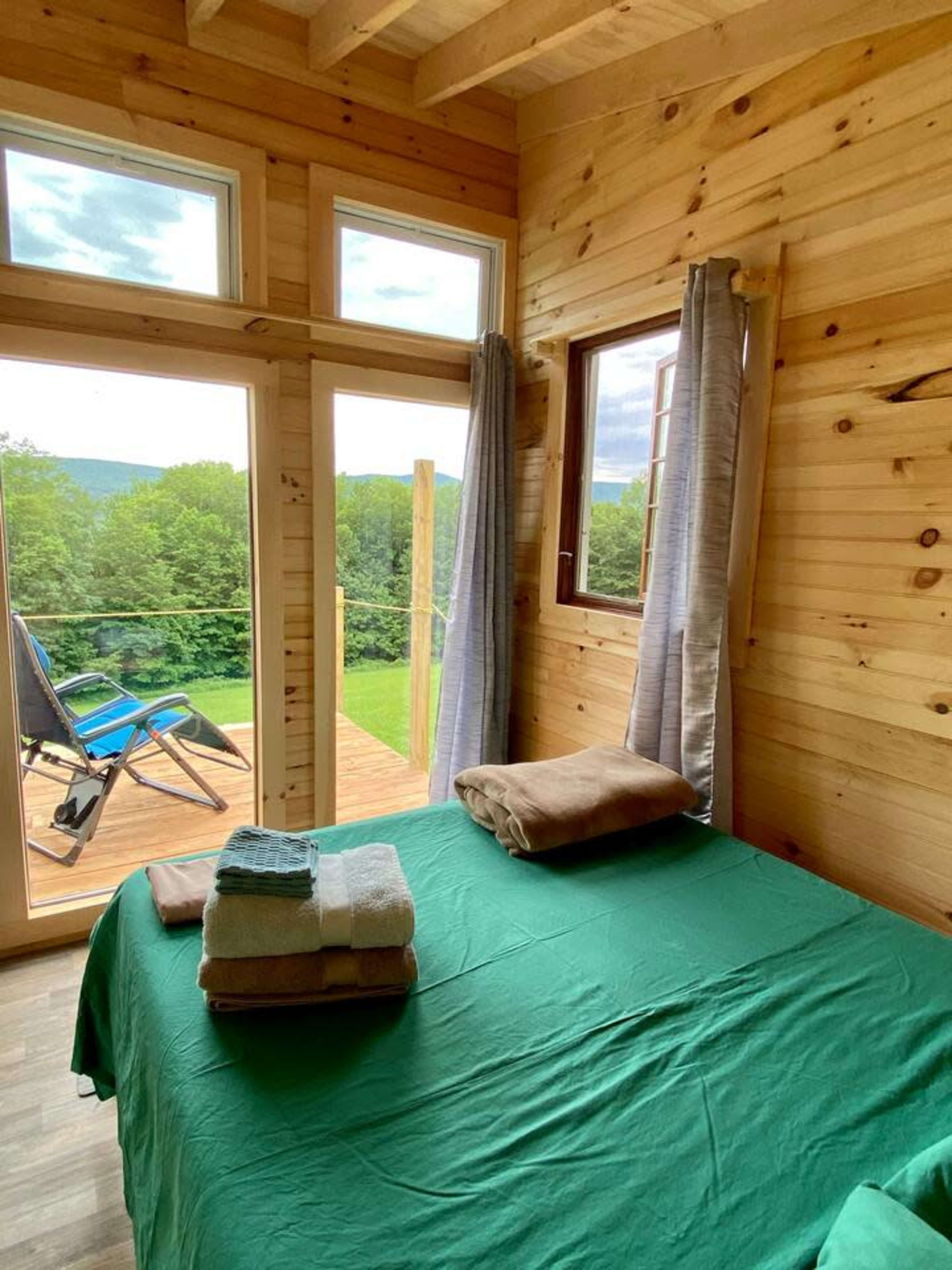 A wooden-paneled room features a bed with neatly arranged towels and large windows opening to a view of greenery and a deck outside.
